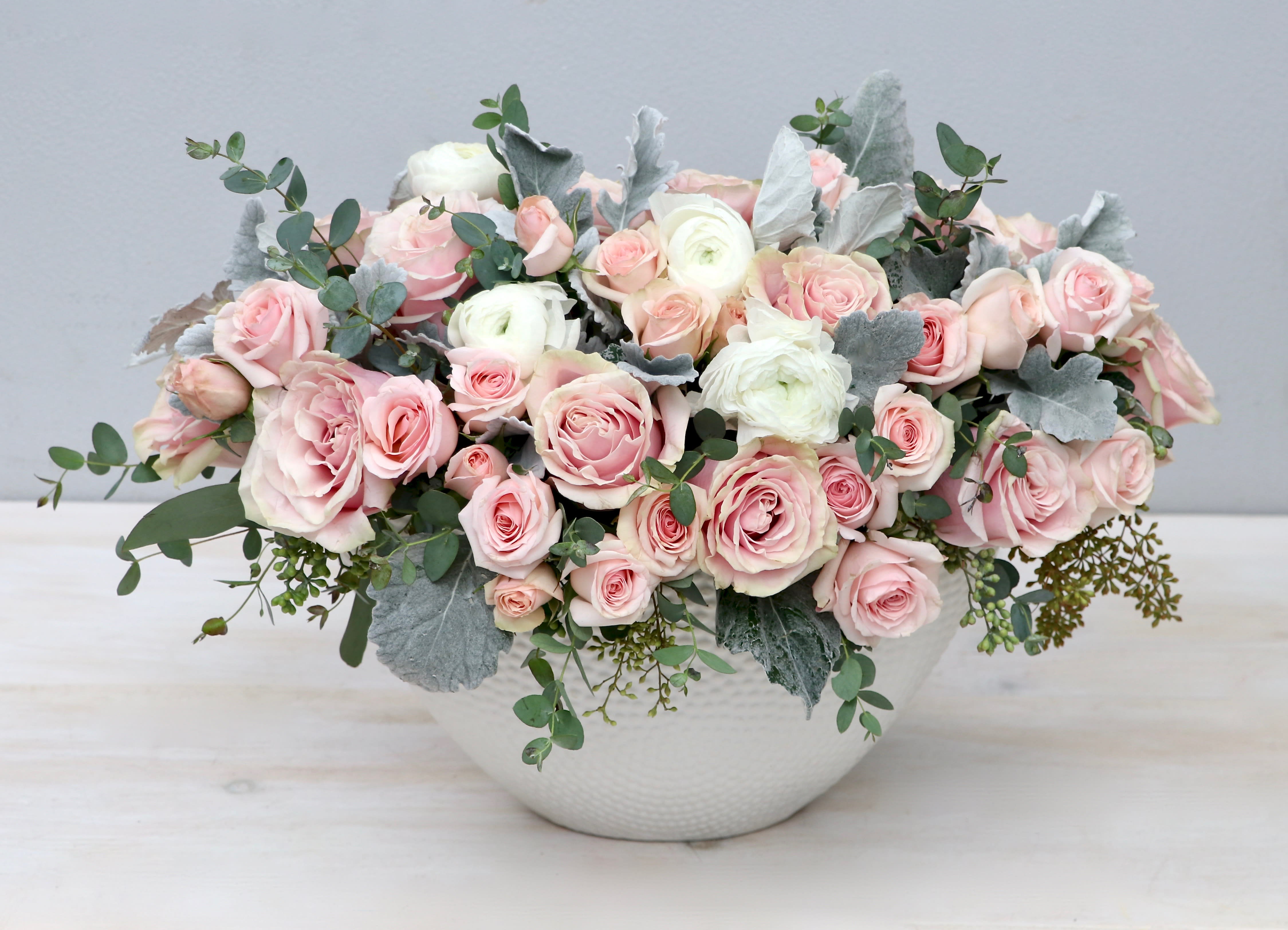 Low arrangement of blush pink roses and white blooms in a white bowl with silver foliage accents.