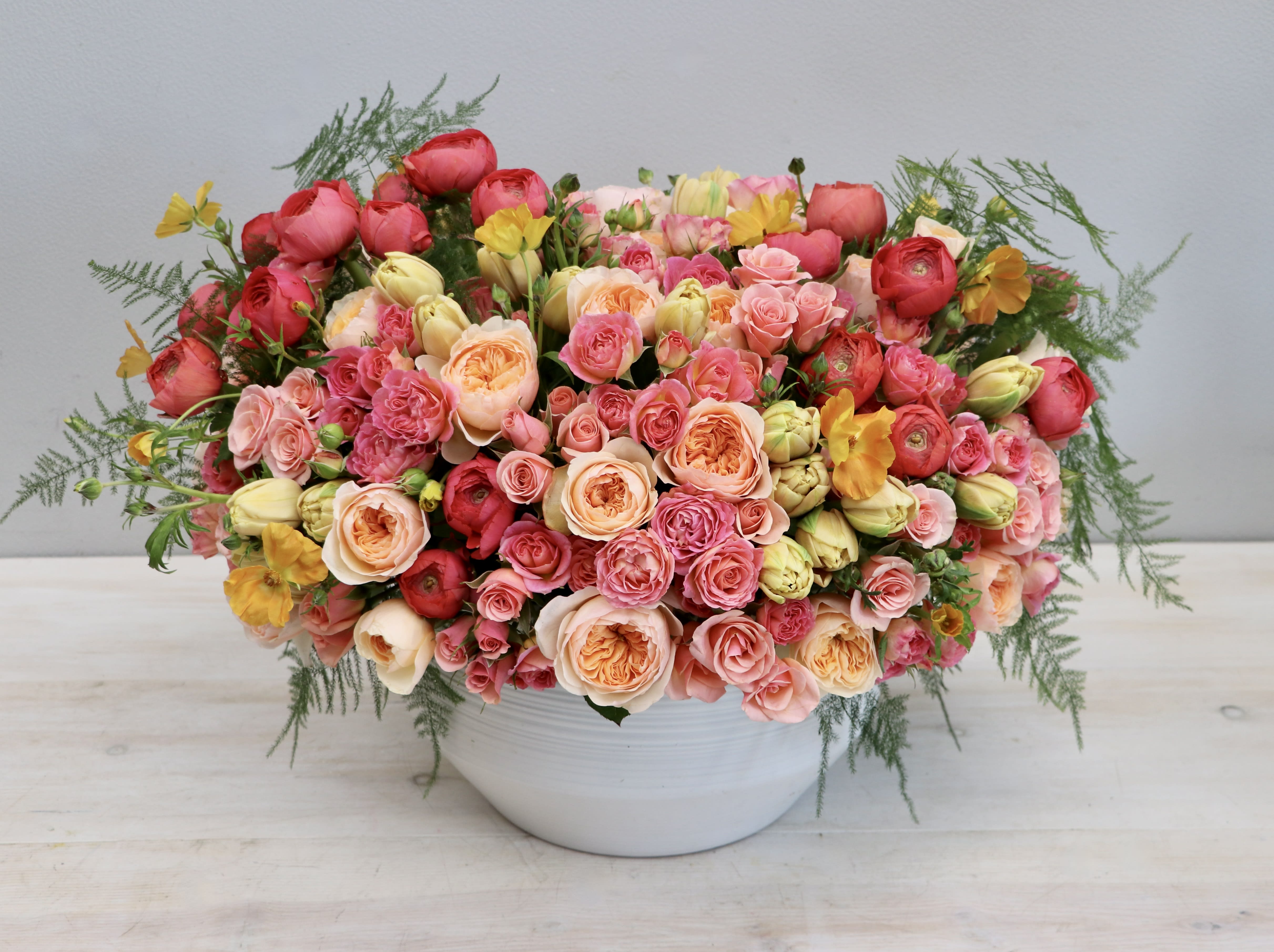 Low white bowl filled with dense pink, peach, and yellow roses and buds in a rounded arrangement.