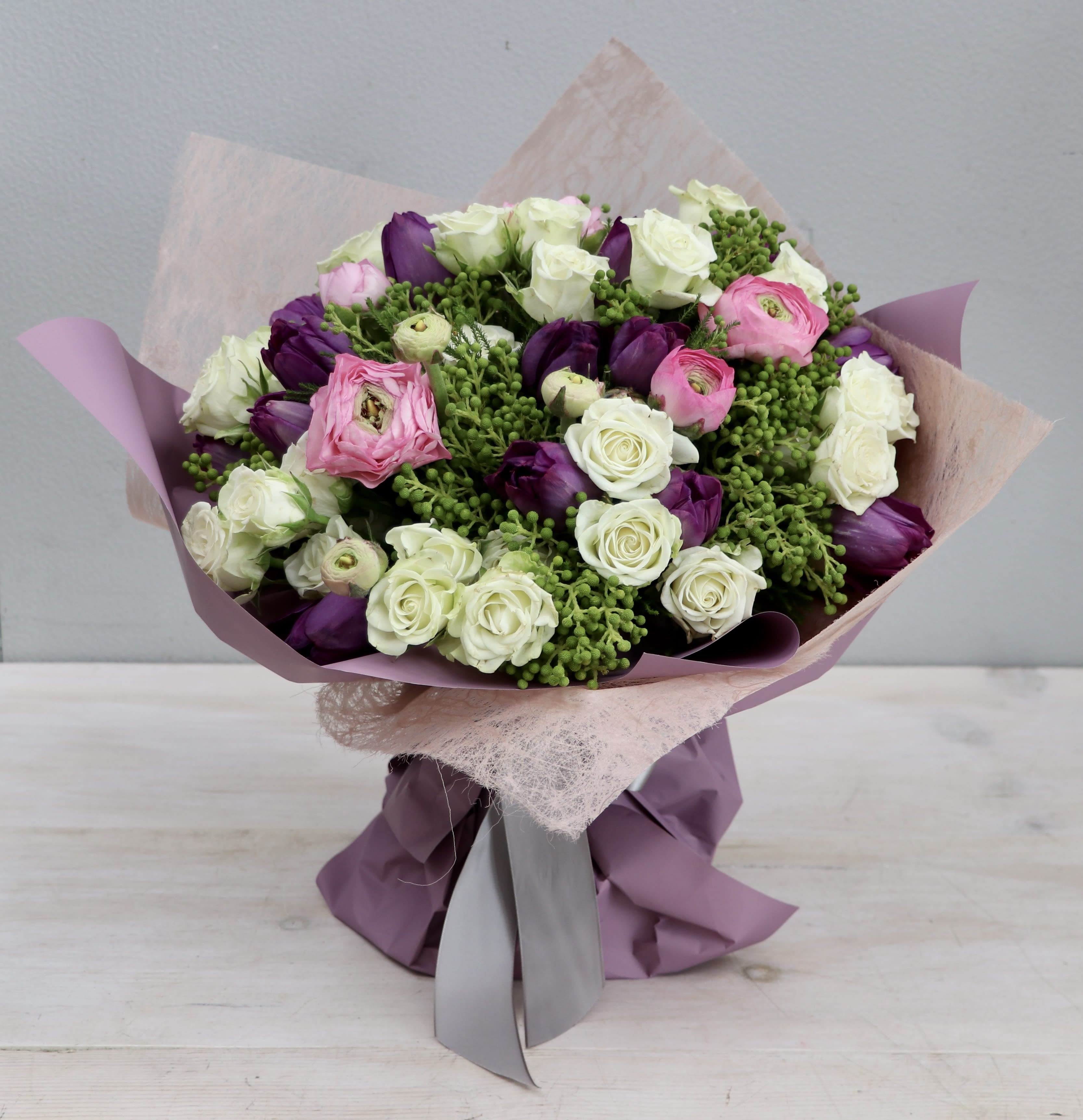 Hand-tied bouquet of white roses, purple tulips, pink ranunculus and green berries in mauve wrapping