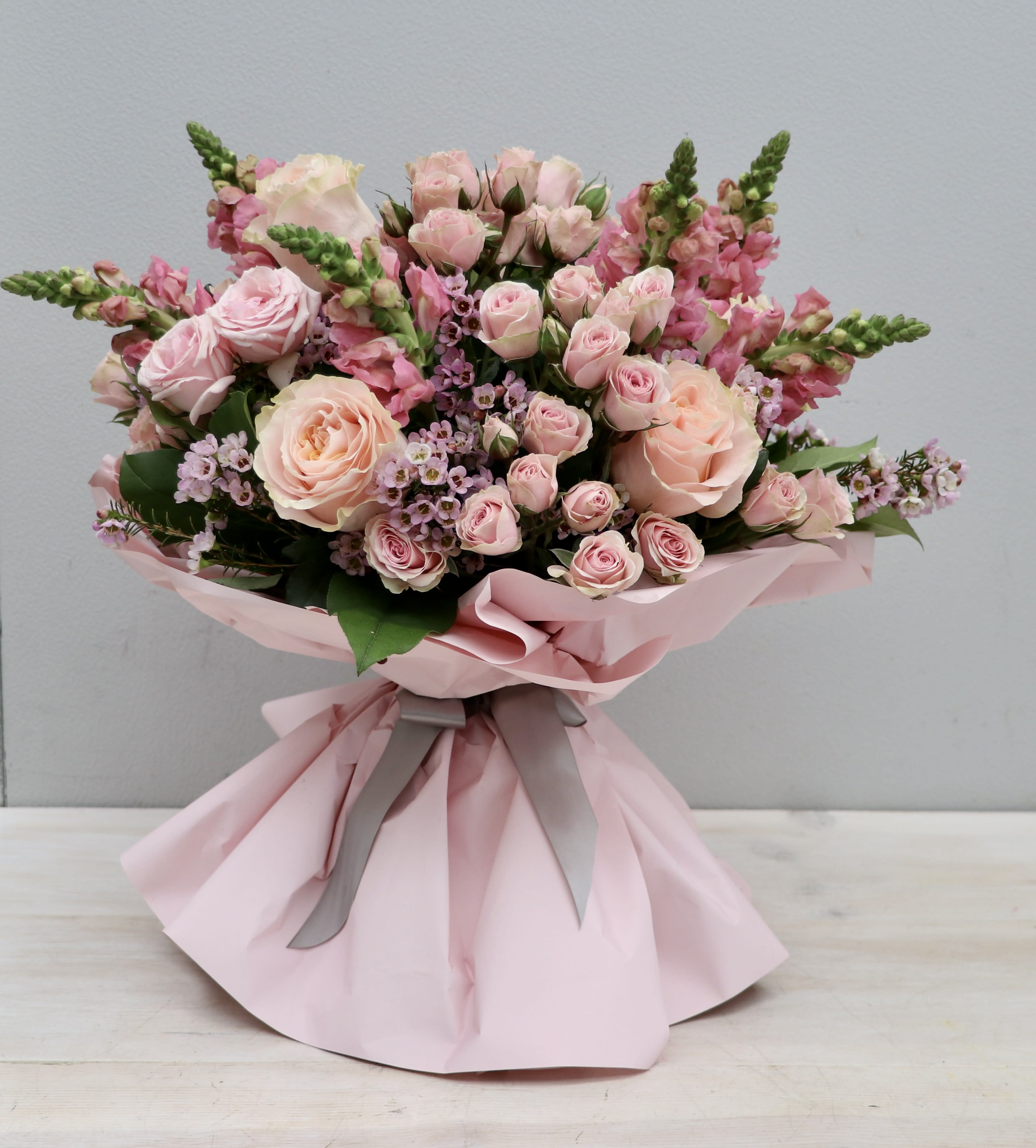 Hand-tied bouquet of light pink roses and snapdragons wrapped in pink paper with gray ribbon