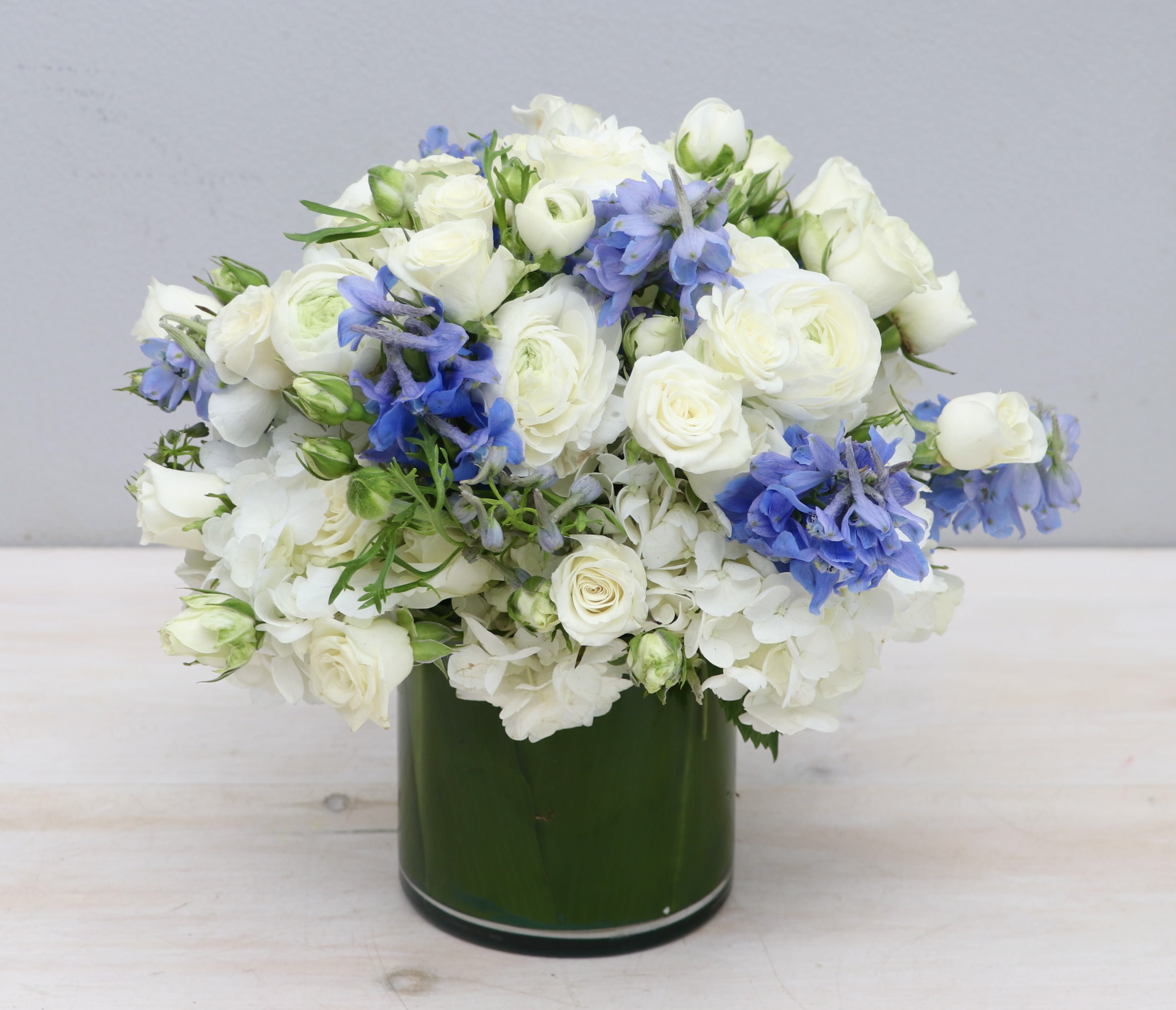 Low round arrangement of white flowers with blue accents in a green cylinder vase