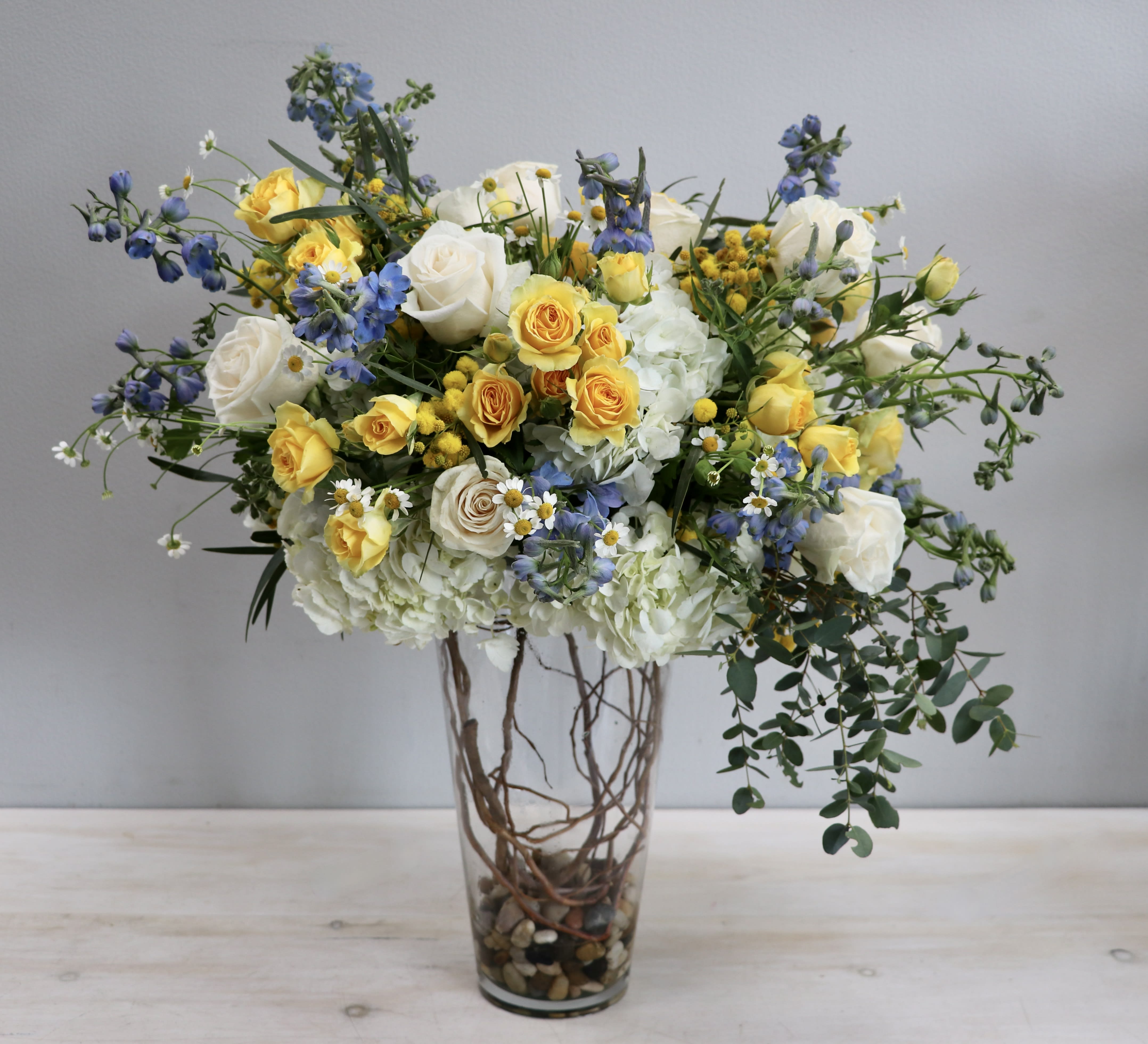 Tall arrangement of yellow and white roses, blue flowers, and hydrangeas in a clear glass vase.