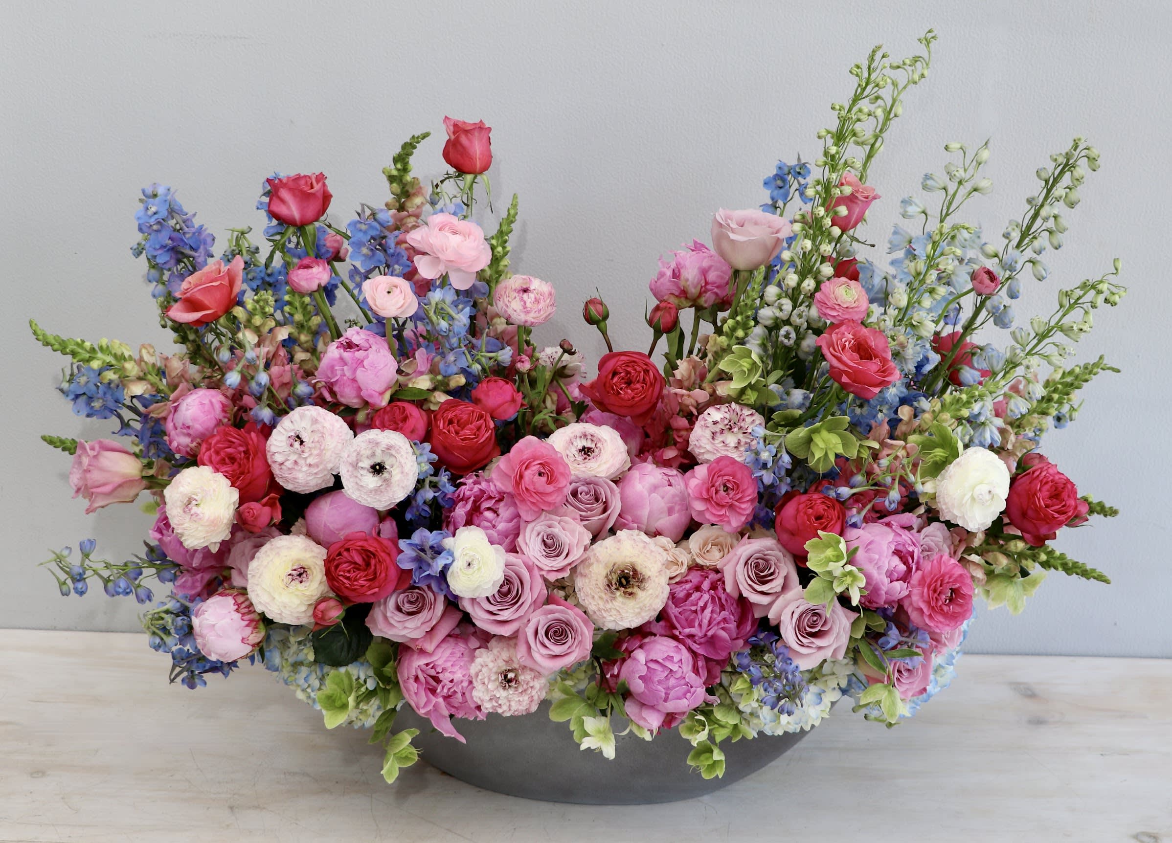Low oval arrangement of pink and red roses, ranunculus, and peonies with blue delphinium in a gray bowl.