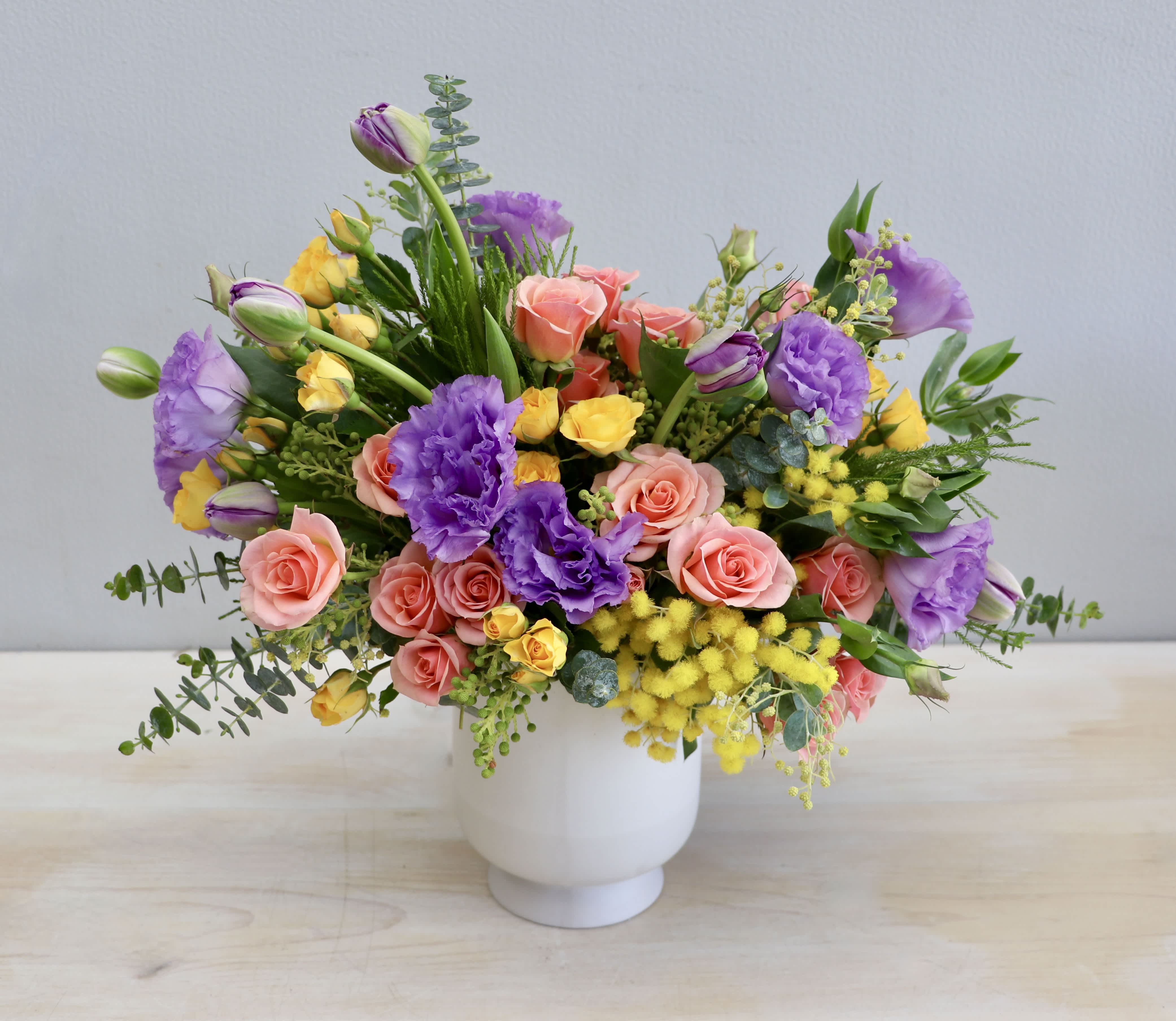 Colorful arrangement of purple lisianthus, peach roses, and yellow blooms in a white ceramic vase