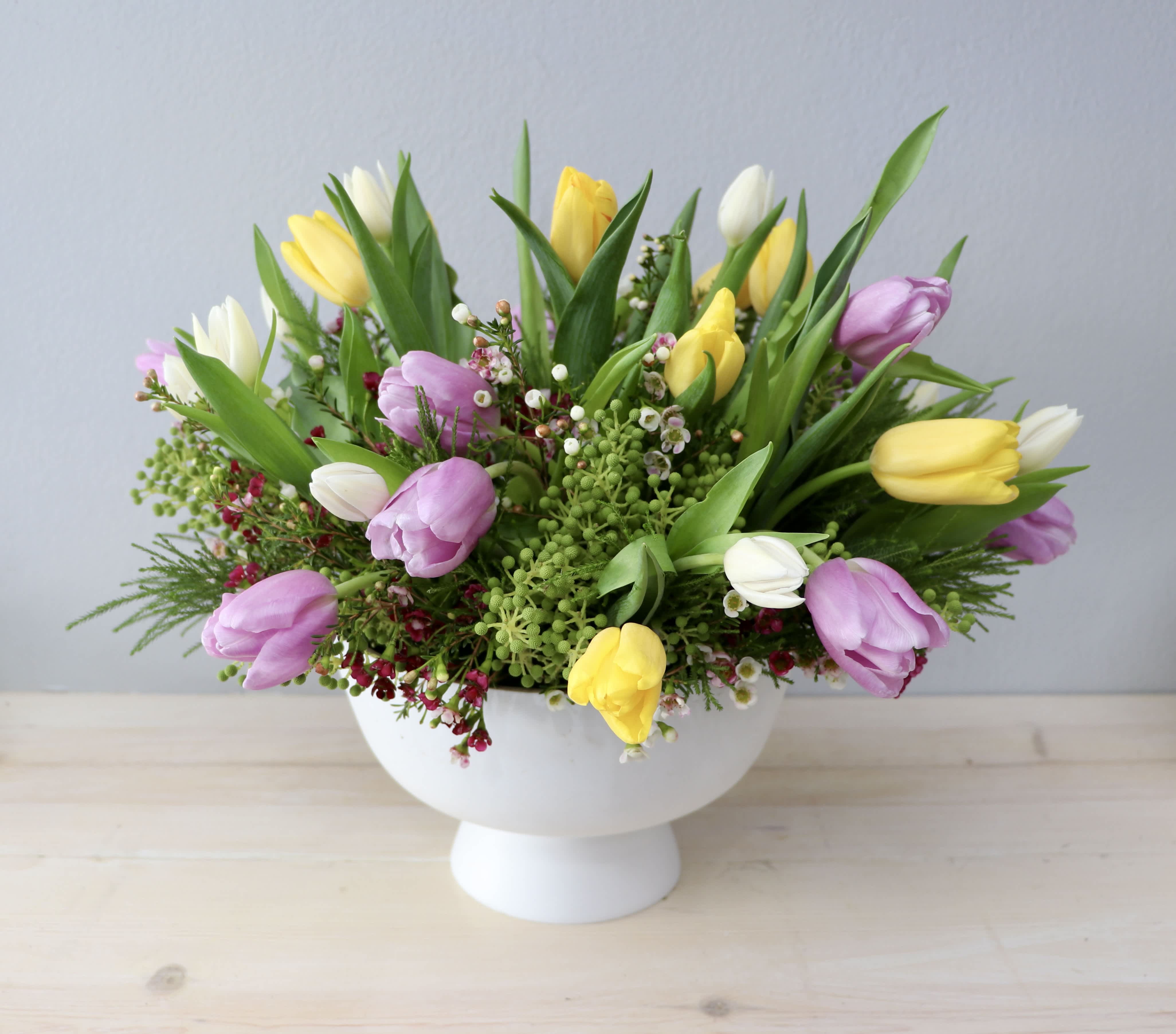 Low arrangement of yellow, white, and lavender tulips in a white pedestal bowl