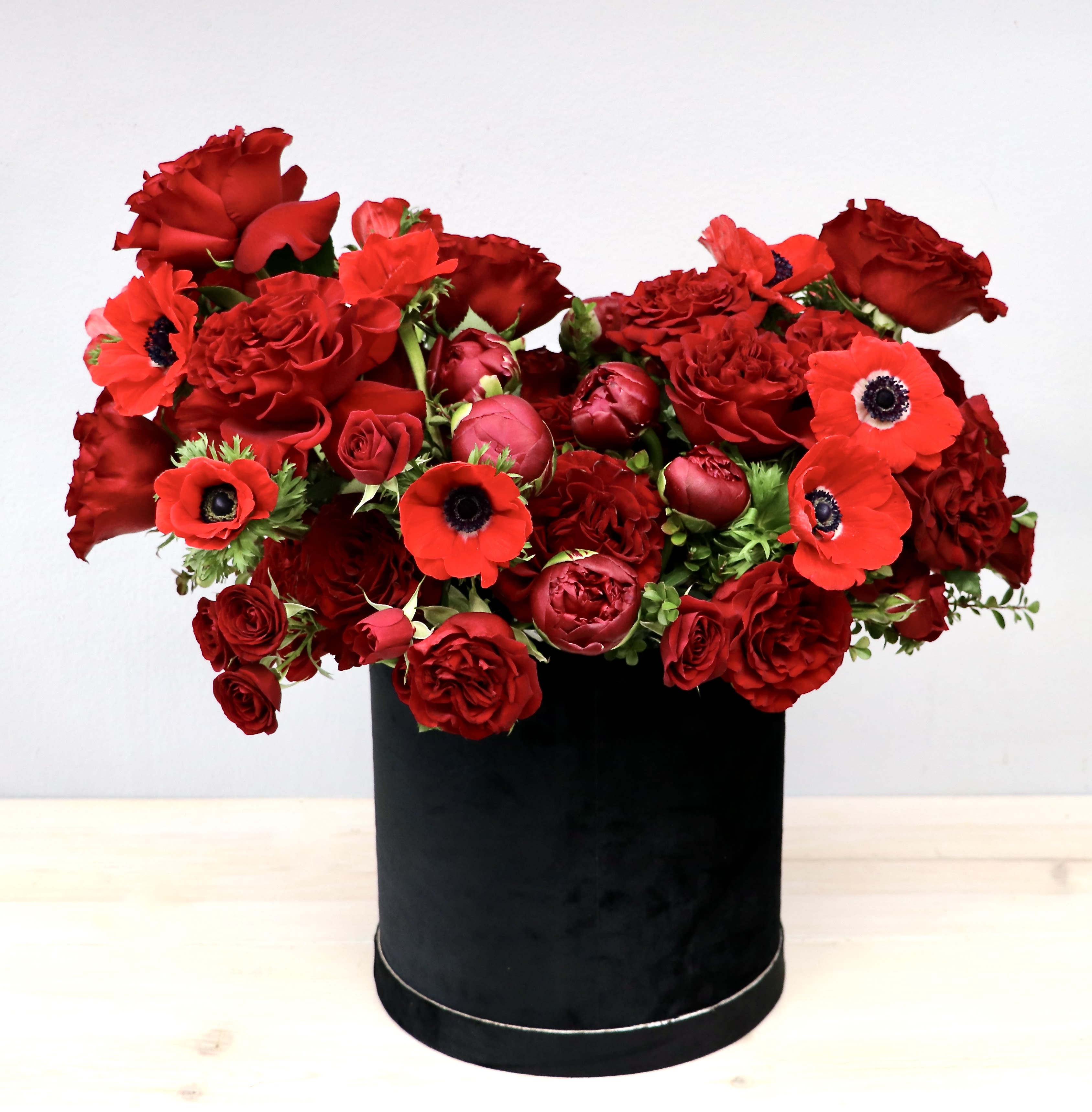 Lush arrangement of red roses, ranunculus, and anemones in a black round hatbox container