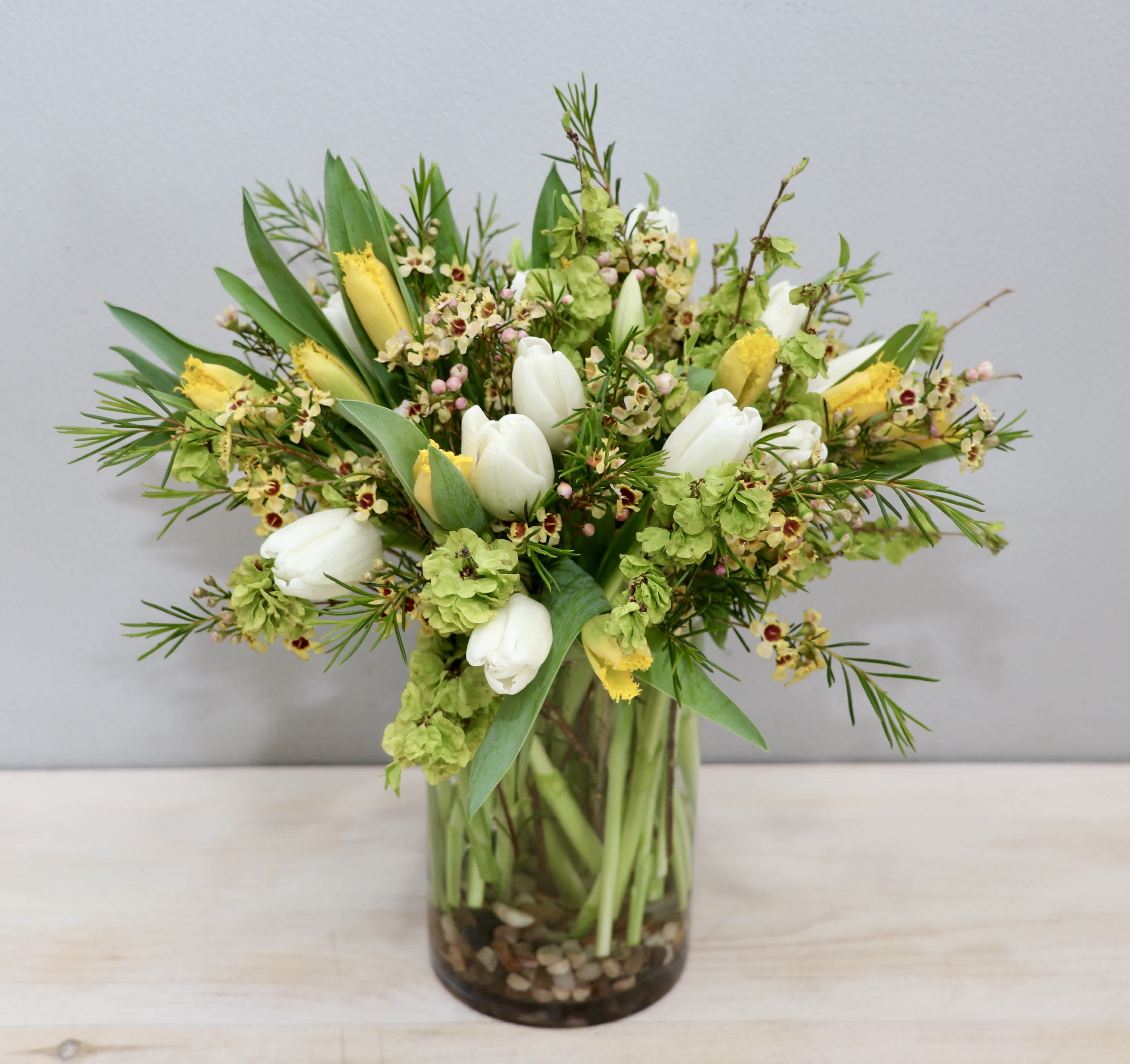 Springy arrangement of white tulips and yellow flowers in a glass cylinder vase with pebbles