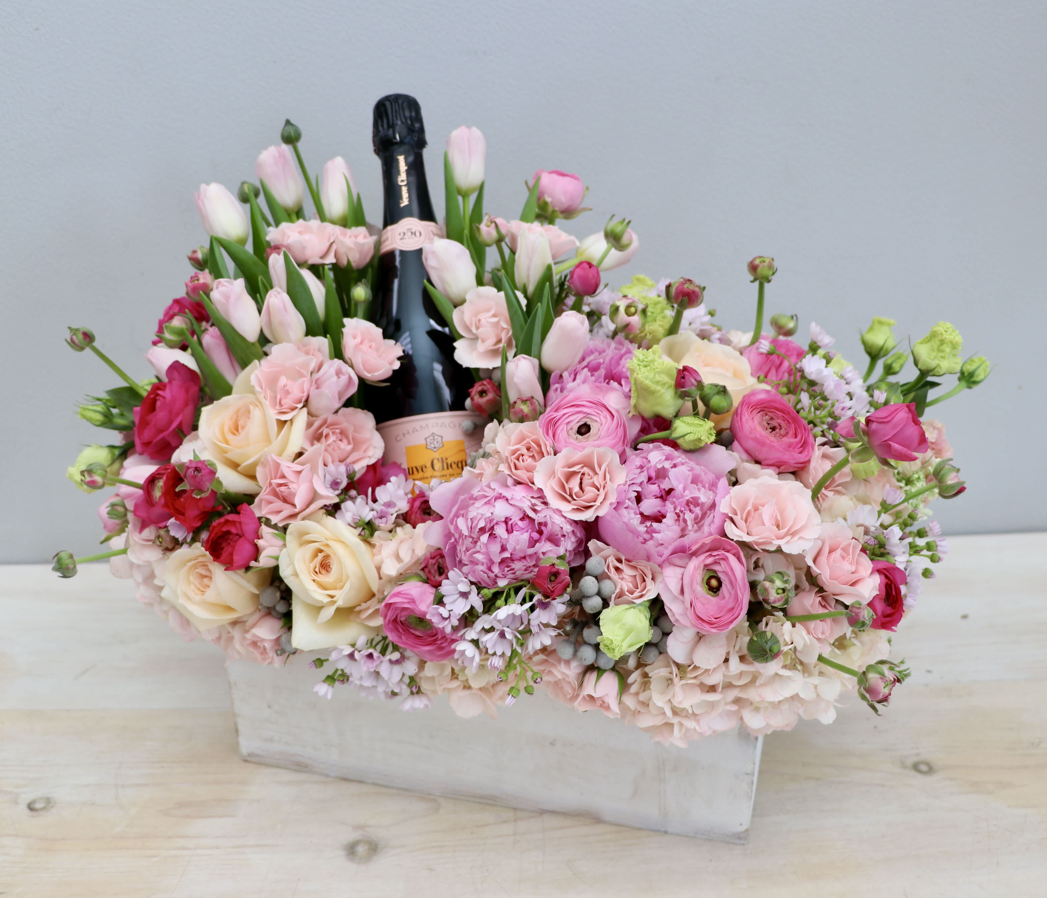 Pink and cream flower arrangement in a white box with a champagne bottle in the center