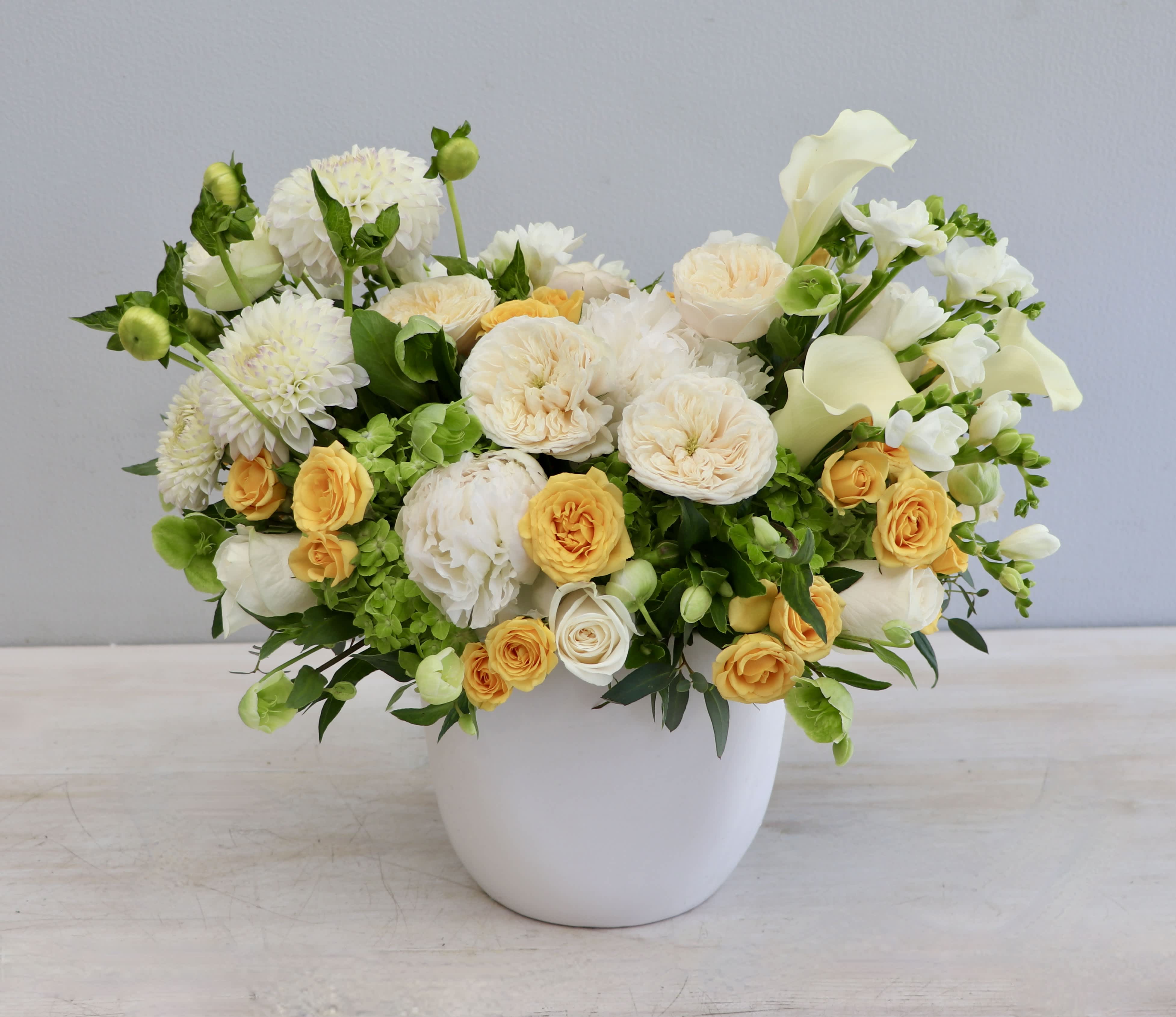 Low arrangement of white and yellow roses, dahlias, and calla lilies in a round white ceramic pot