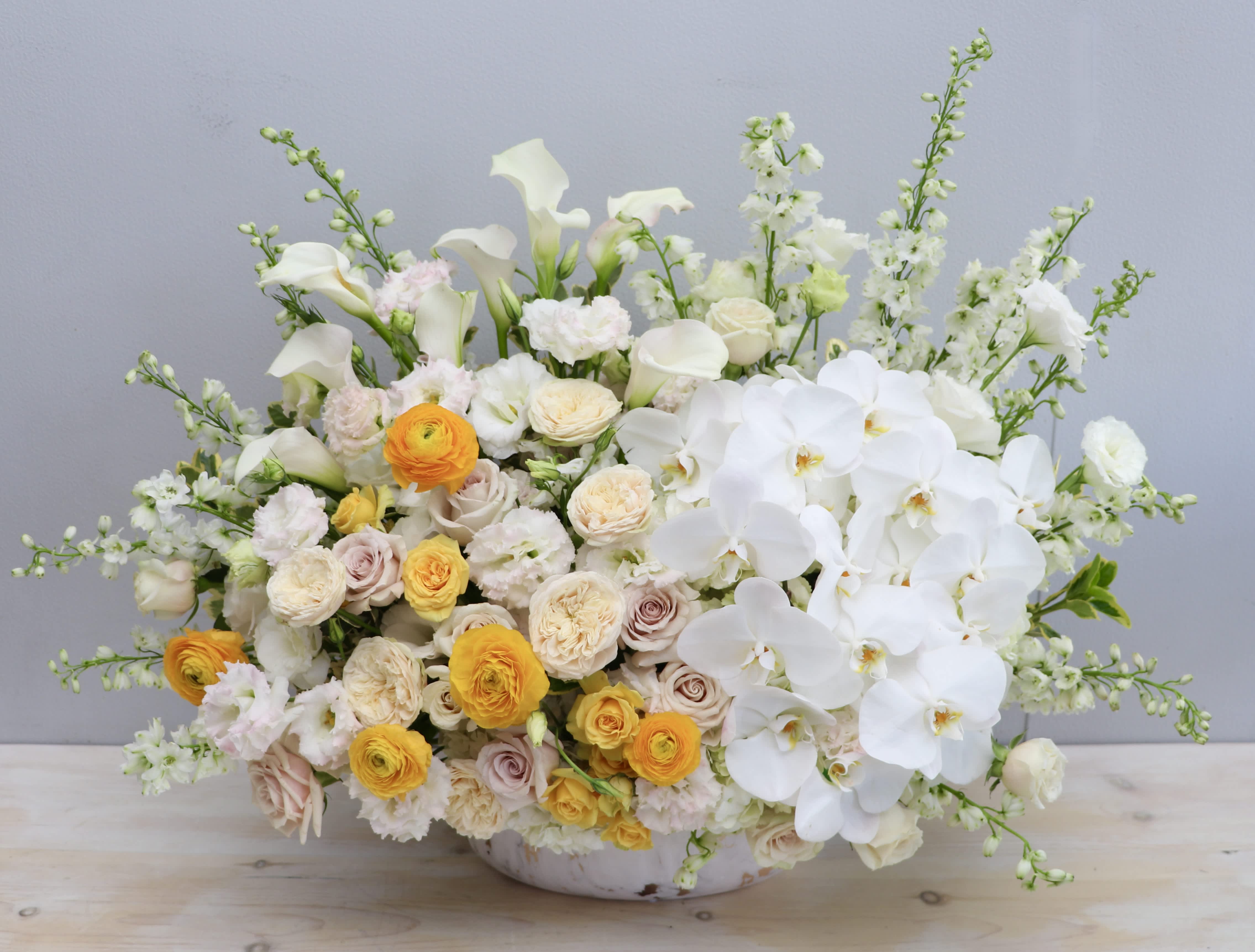 Large low arrangement of white orchids, calla lilies, and yellow and cream roses in a white bowl.