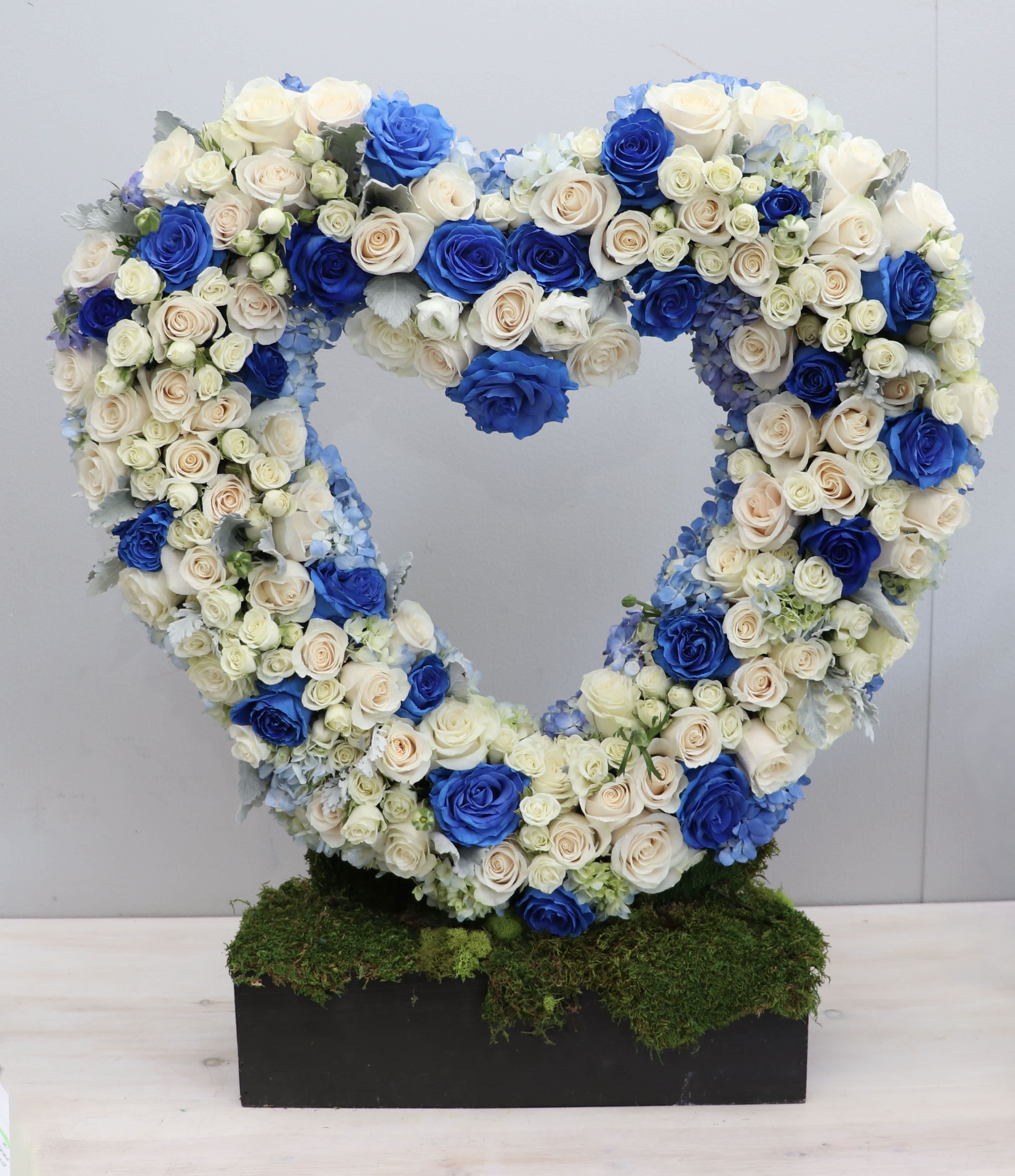 Heart-shaped standing wreath of blue and ivory roses on a moss-covered rectangular base.