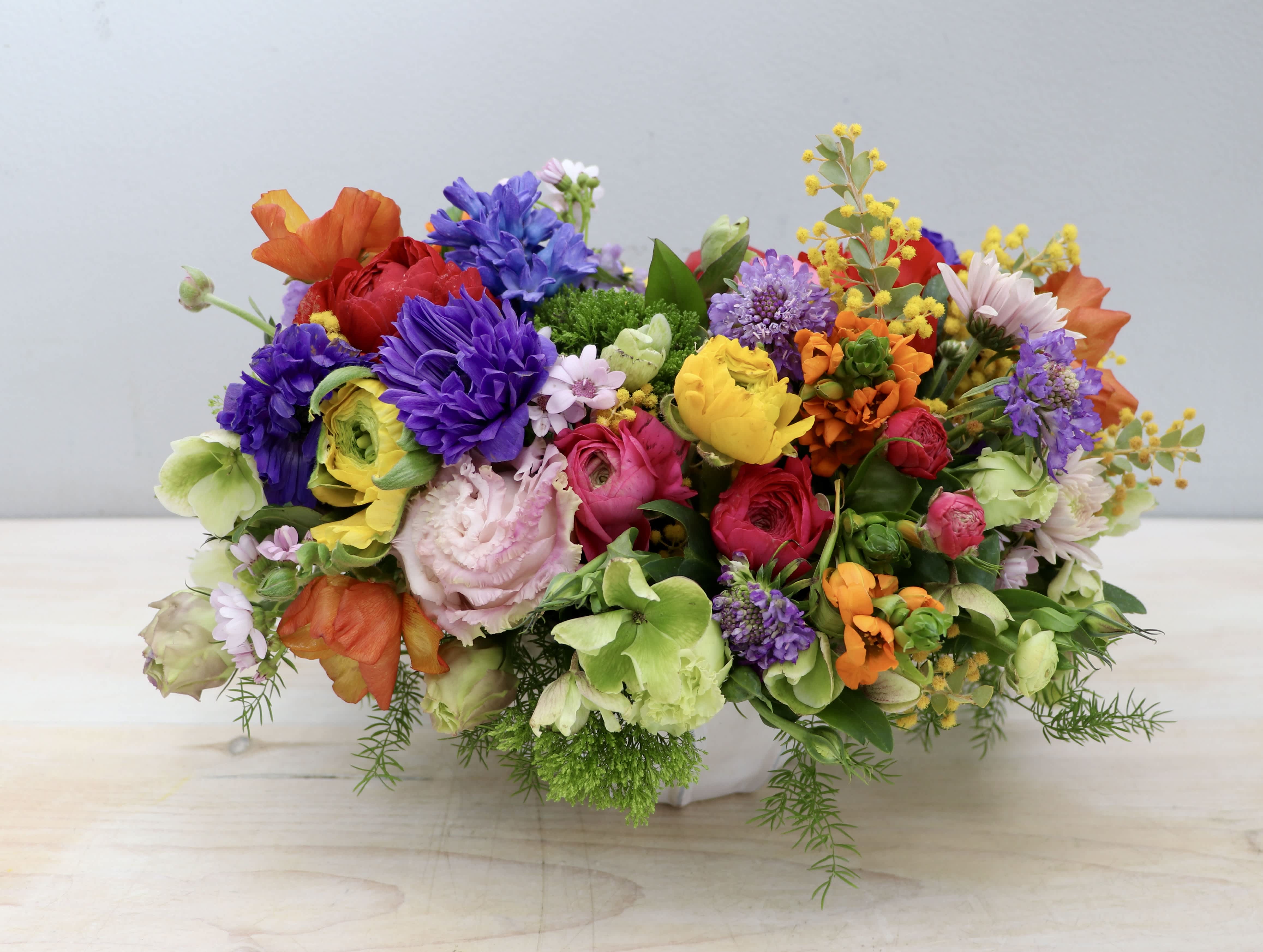 Low, oval arrangement of bright mixed flowers in a white ceramic container on a light wood surface