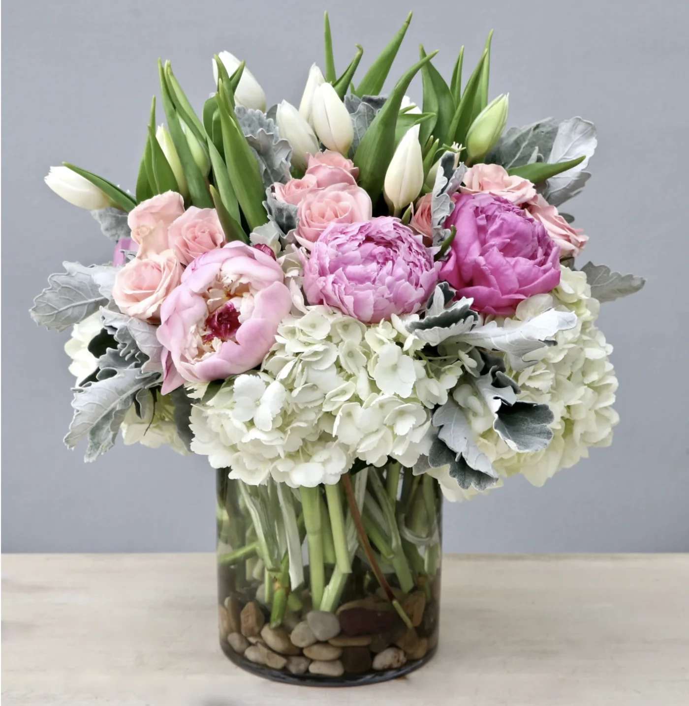 Pink peonies, pale roses, white tulips, and hydrangeas arranged in a clear glass cylinder vase with stones