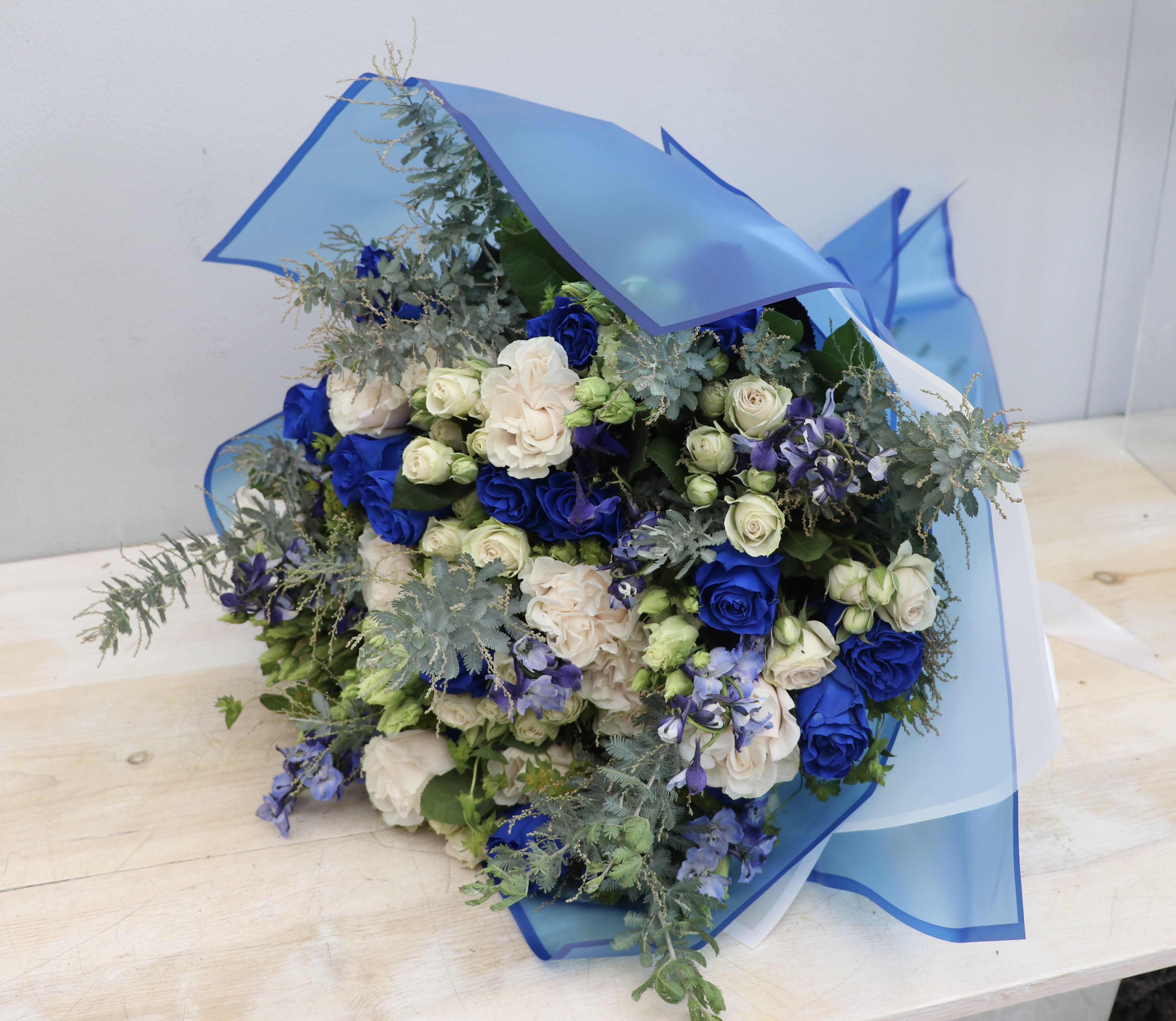 Bouquet of blue and cream roses with small white buds wrapped in blue cellophane.