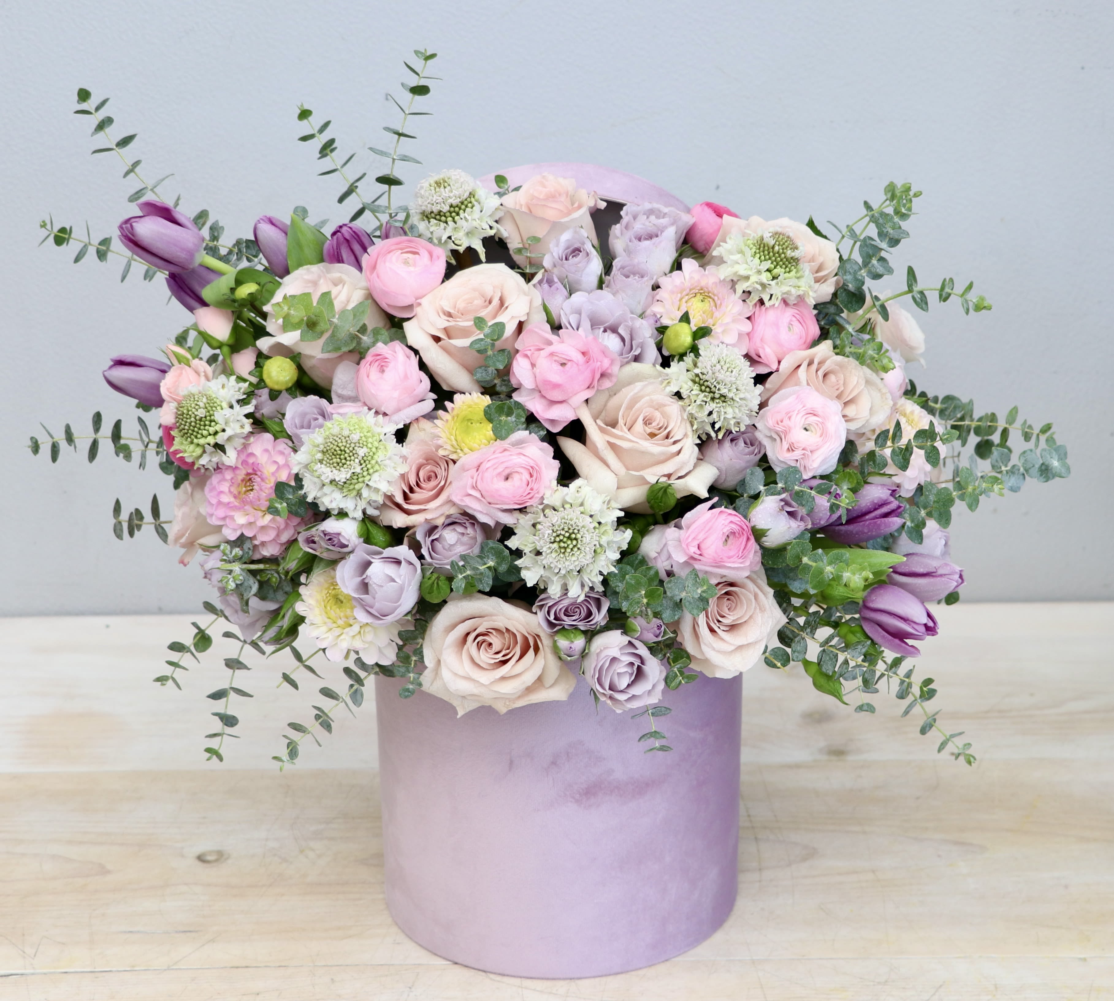 Pastel pink and lavender flower arrangement in a round lavender hatbox-style container
