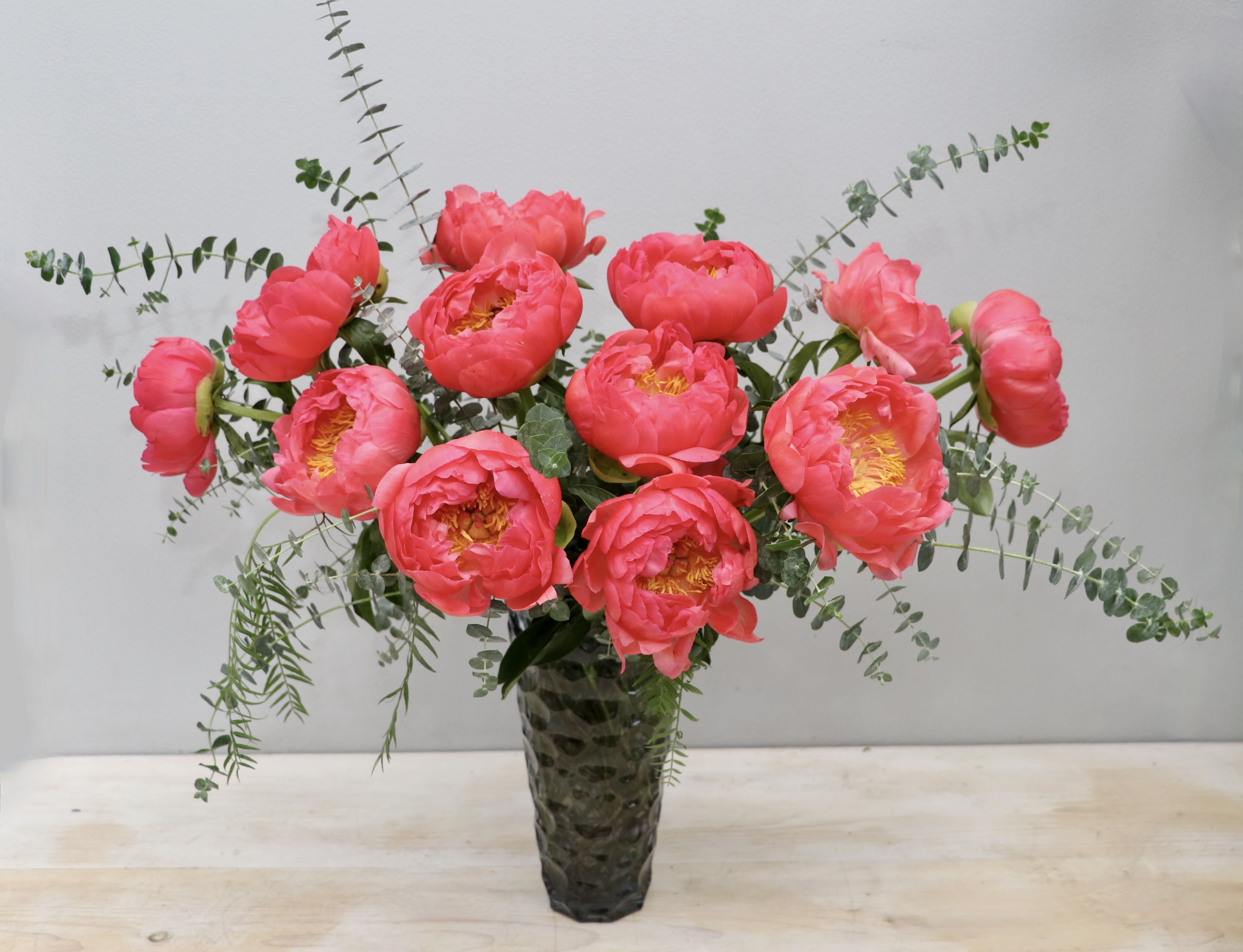 Tall arrangement of bright pink peonies with trailing eucalyptus in a textured glass vase