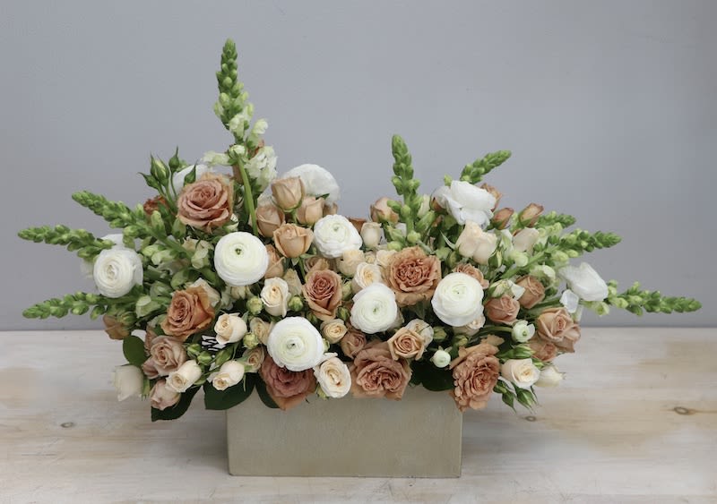 Low rectangular arrangement of white ranunculus and peach roses with snapdragons in a beige container