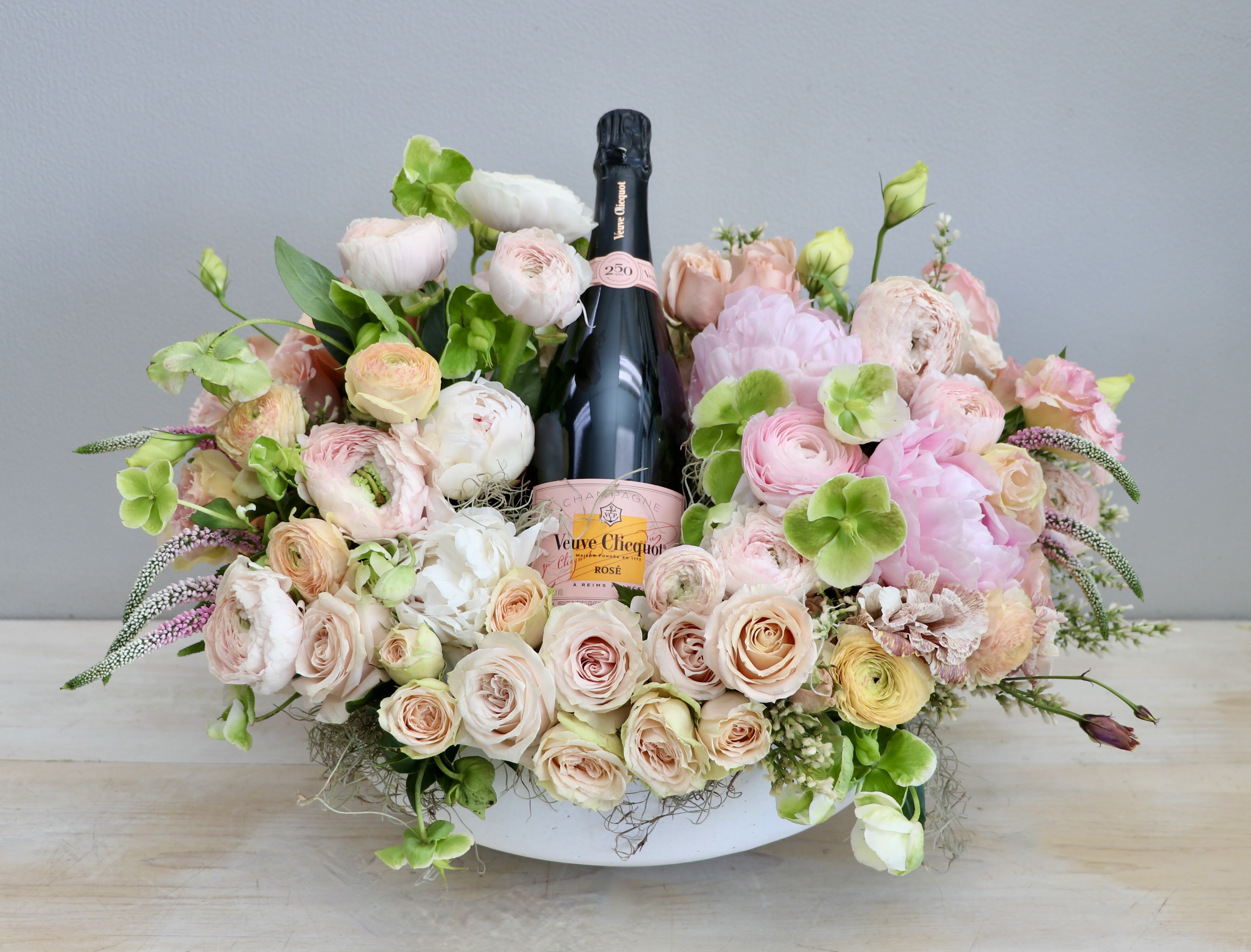 Low pastel flower arrangement in a white bowl with a pink champagne bottle in the center