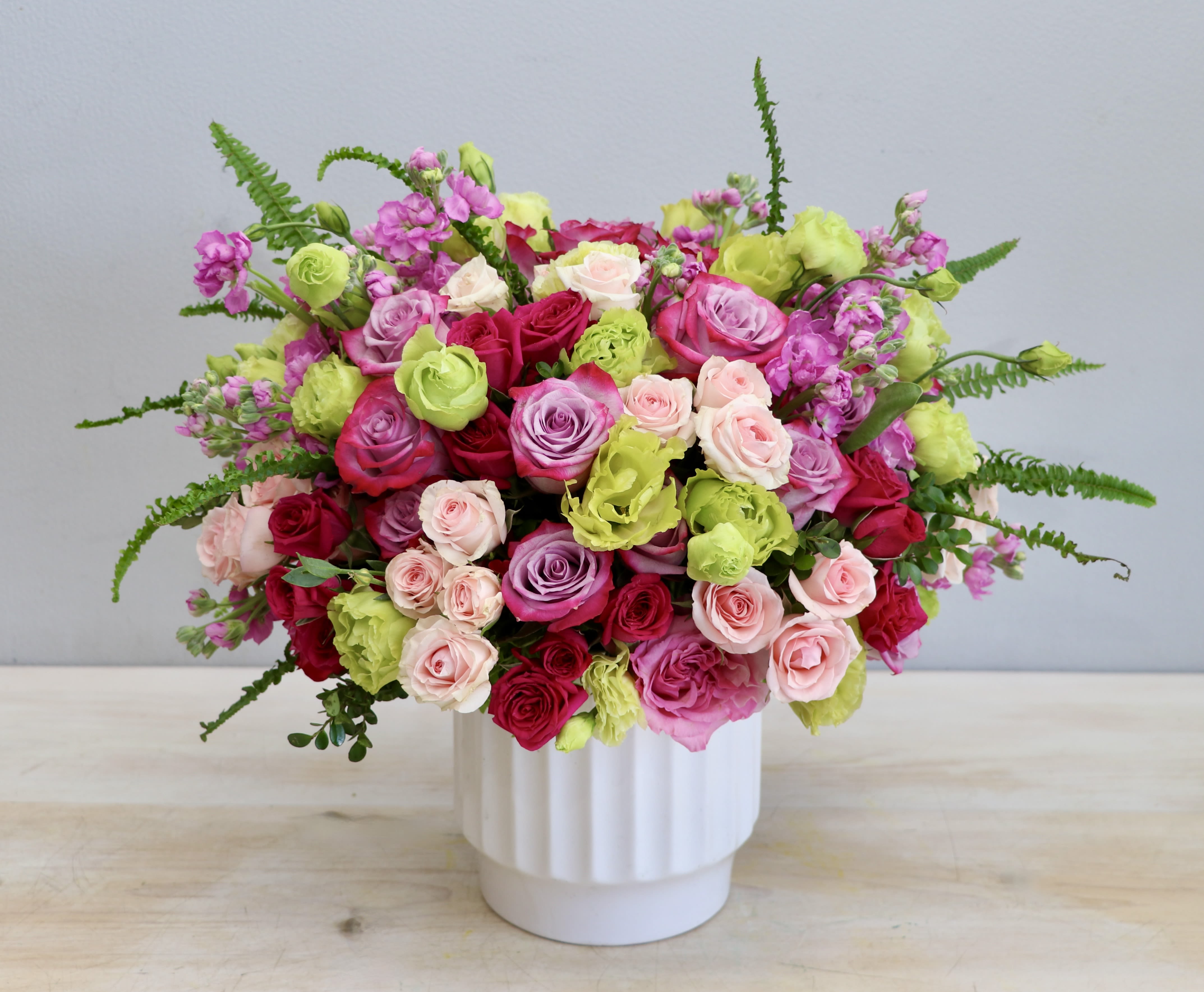 Lush arrangement of pink, red, lavender roses and green blooms in a white vase