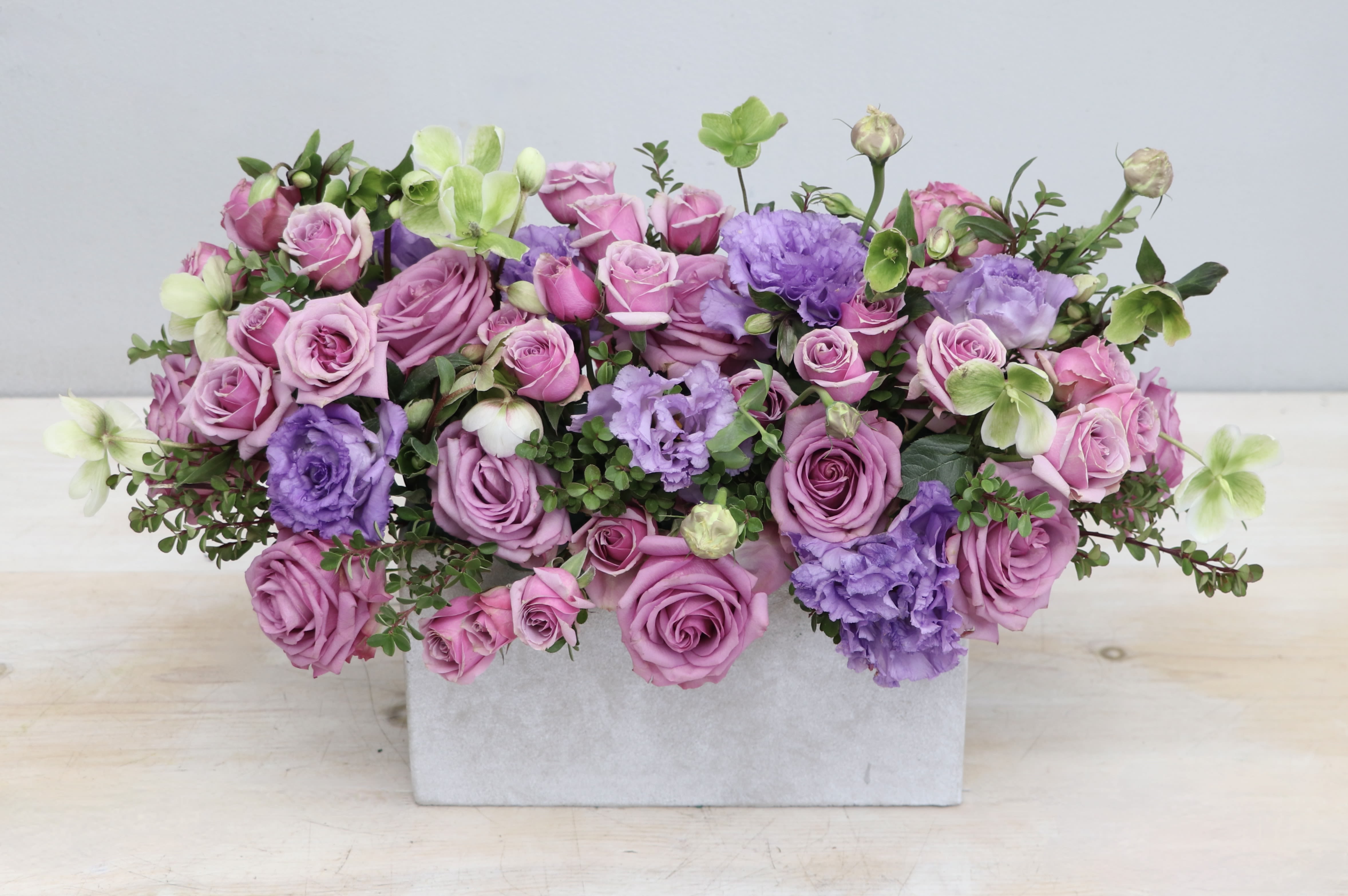 Low arrangement of pink and lavender roses with purple blooms in a rectangular white vase