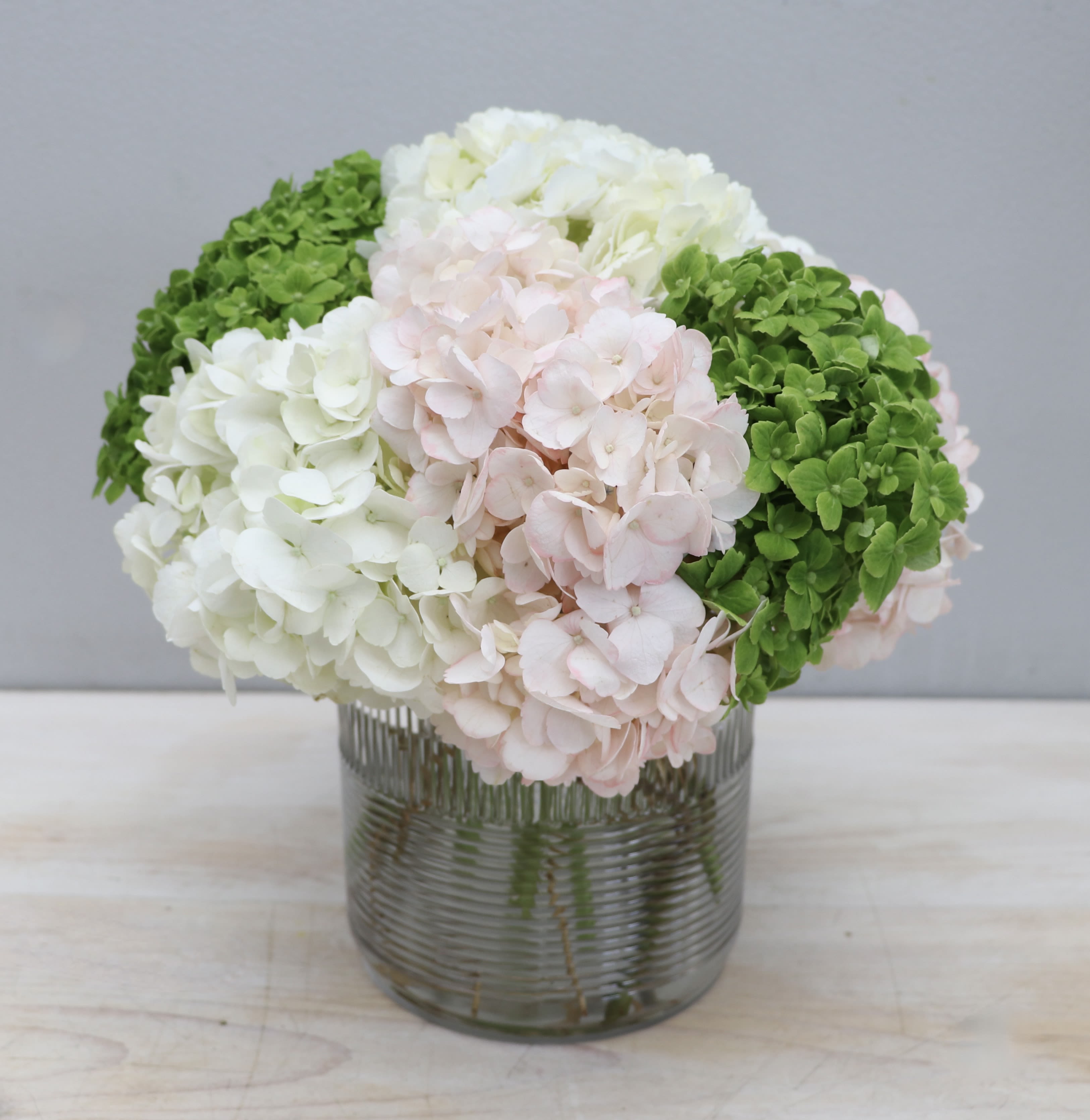 Compact arrangement of white, pale pink, and green hydrangeas in a clear ribbed glass vase