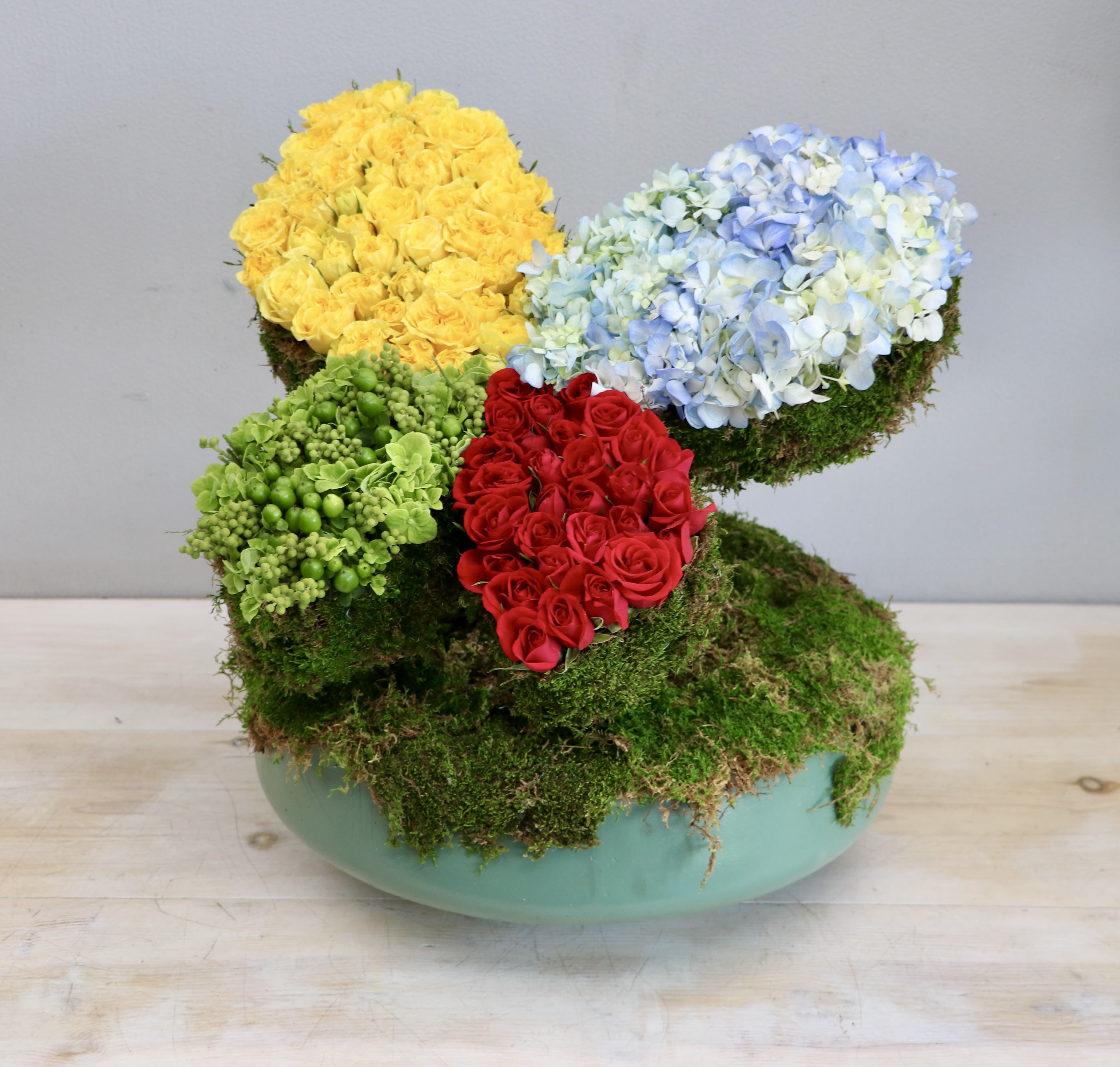 Modern moss-covered arrangement with yellow and red roses and blue hydrangeas in a low green bowl