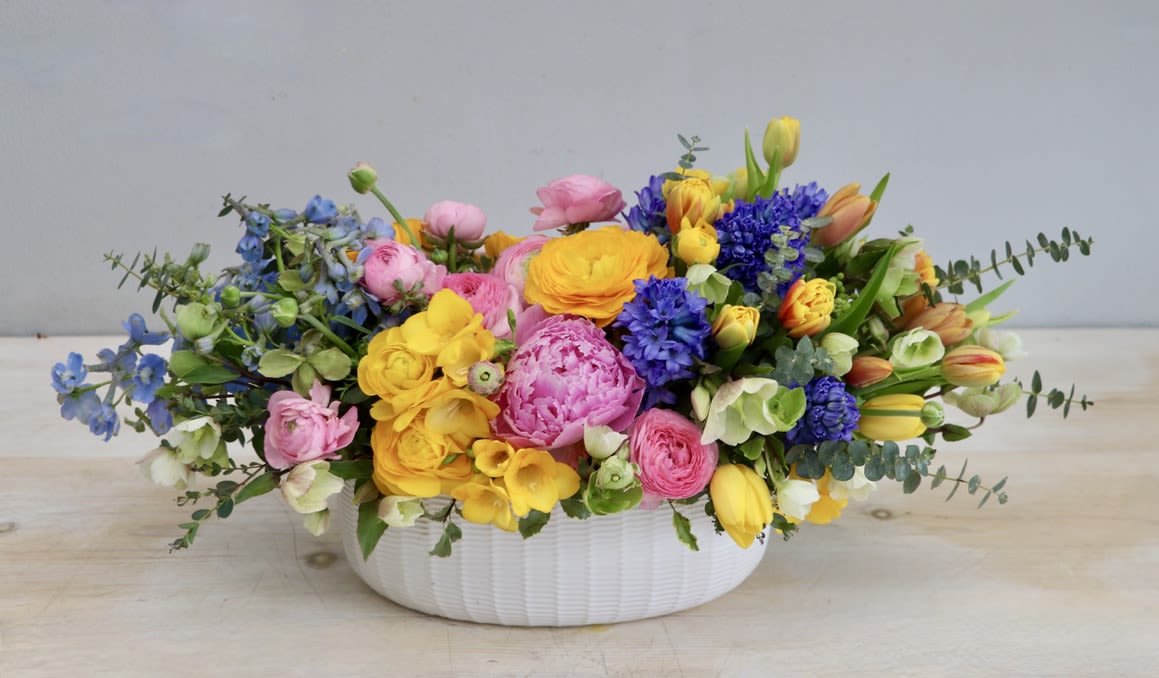 Low white bowl of bright pink, yellow, orange, and blue spring flowers in a lush arrangement