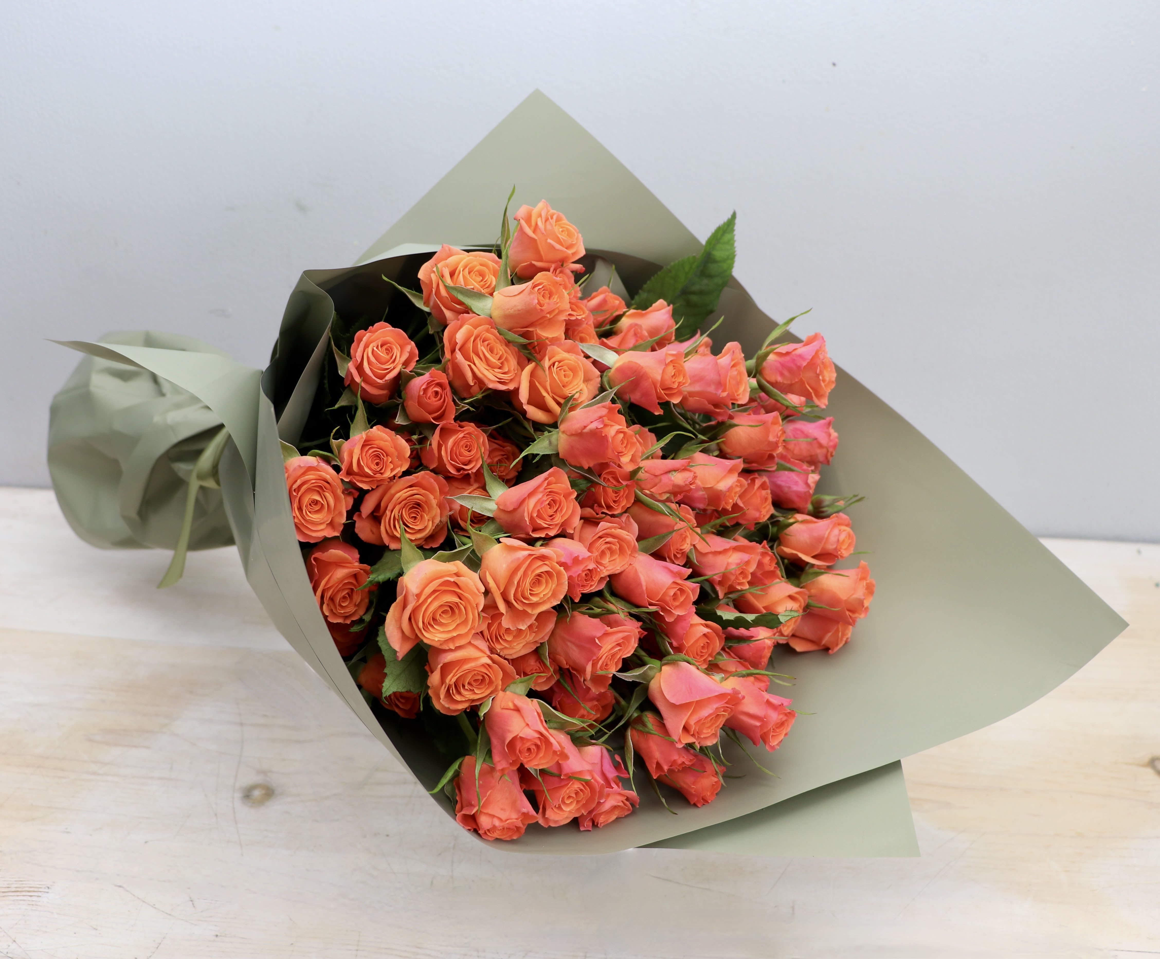 Bouquet of bright orange roses wrapped in matte sage green paper, resting on a light wooden surface