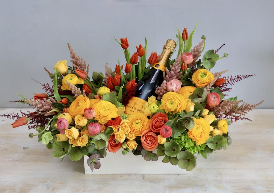 Rectangular arrangement of yellow and orange flowers with red tulips surrounding a bottle of champagne.