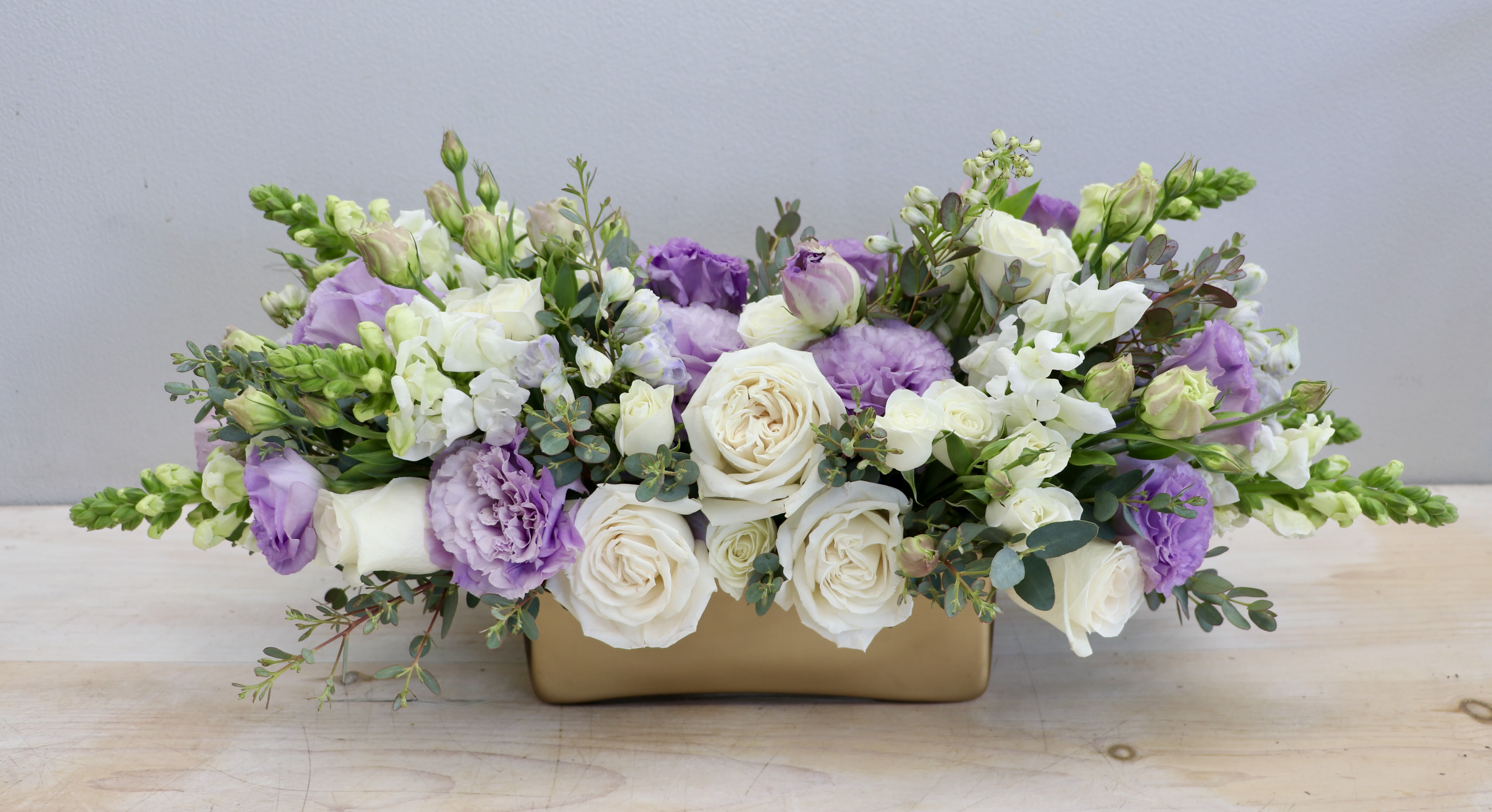 Low arrangement of white roses, purple lisianthus, and white snapdragons in a rectangular gold container.