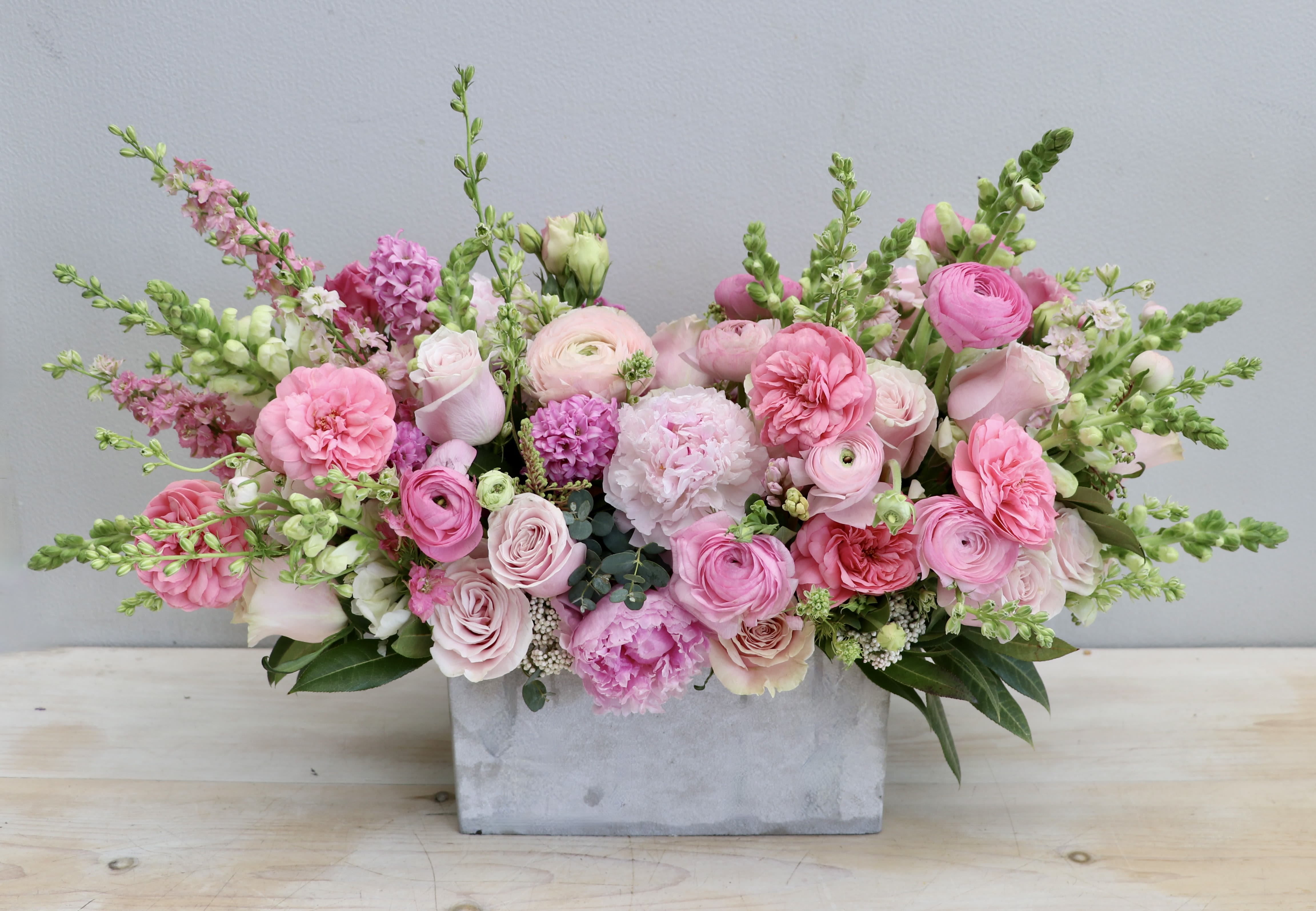 Low rectangular centerpiece of pink roses, ranunculus, peonies and snapdragons in a concrete container.
