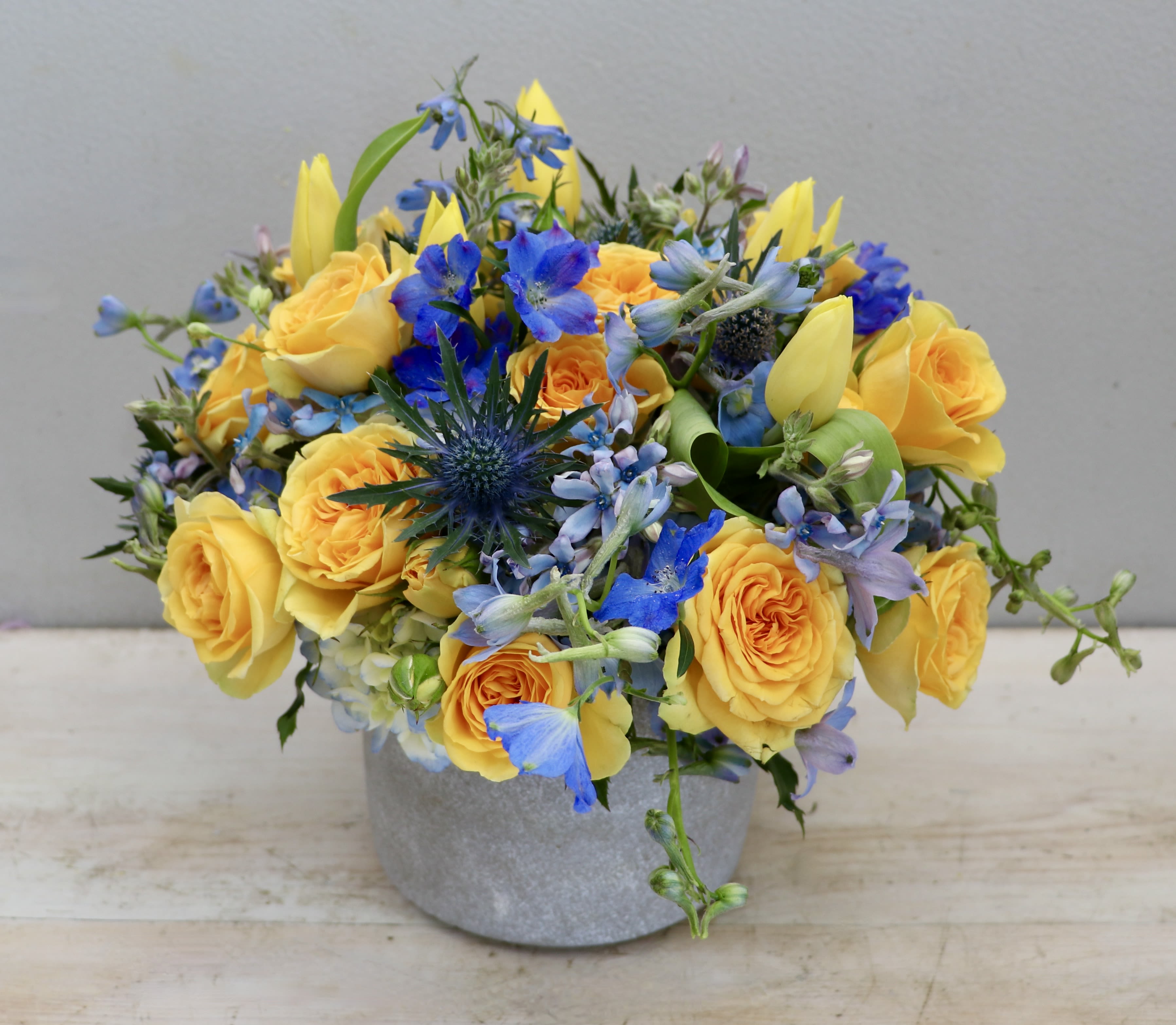 Low arrangement of yellow roses and tulips with blue flowers in a gray pot
