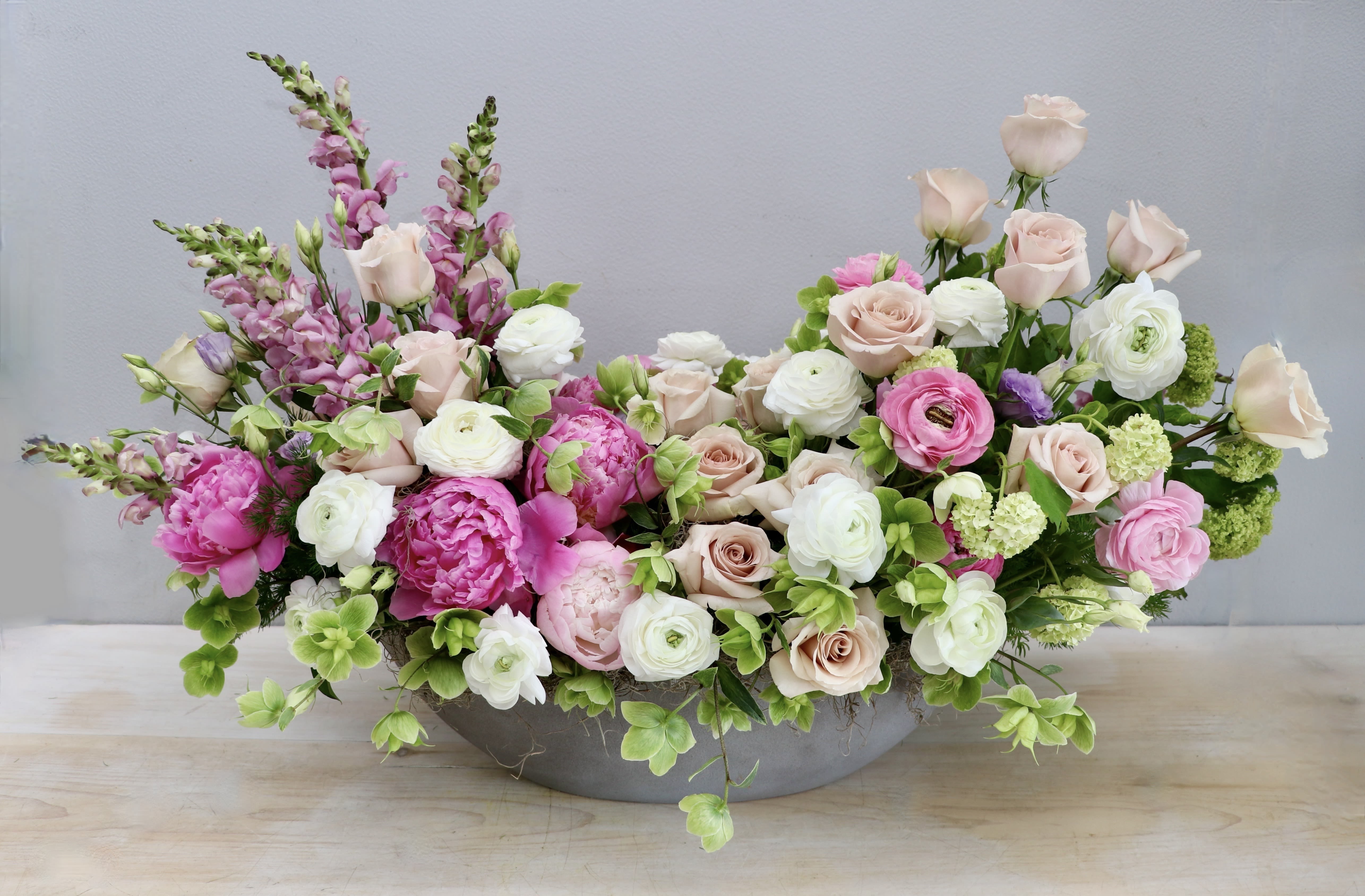 Low oval centerpiece of pink peonies, blush roses and white ranunculus in a gray container
