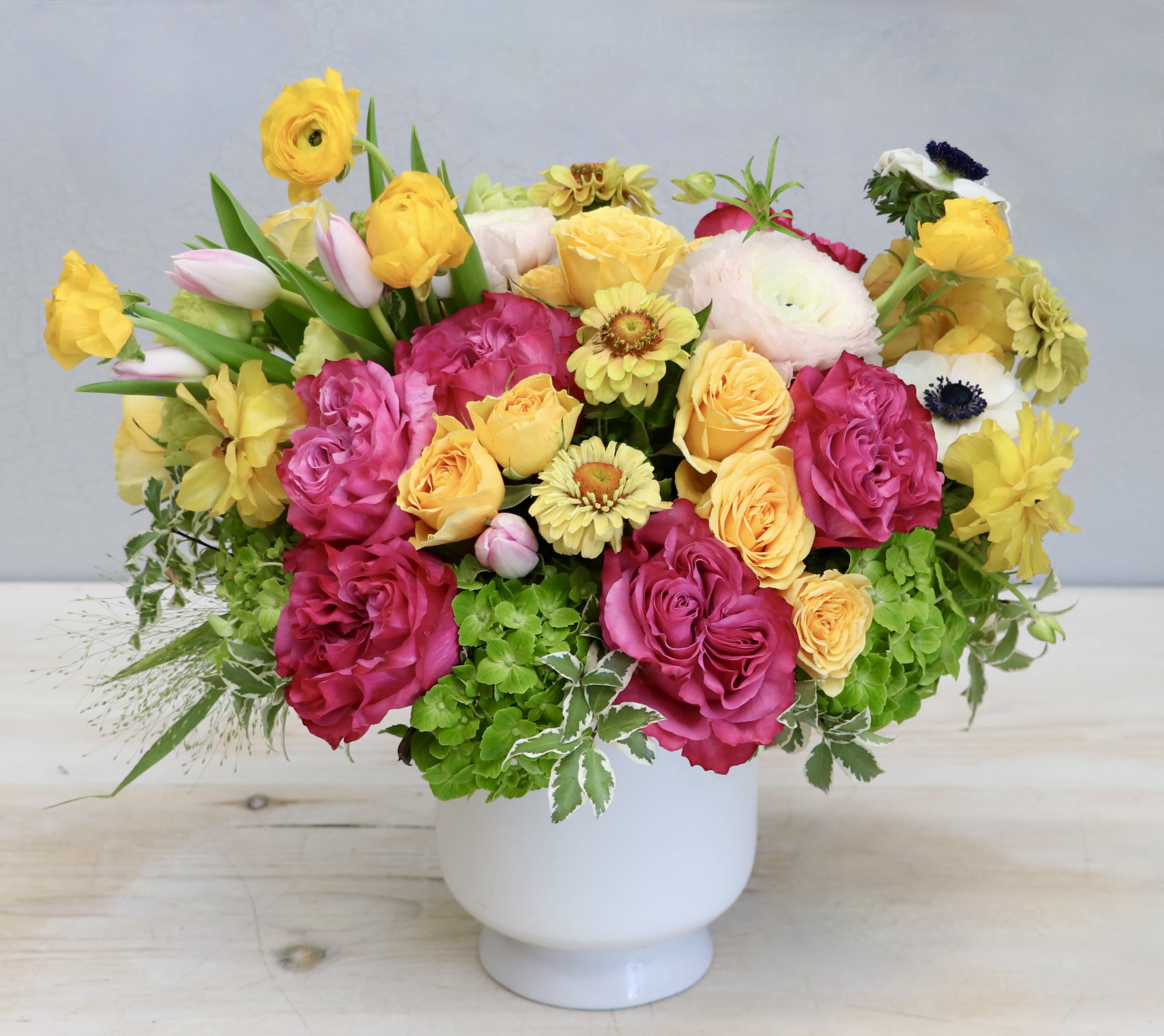 Bright arrangement of pink roses, yellow blooms, and green hydrangea in a white pedestal vase