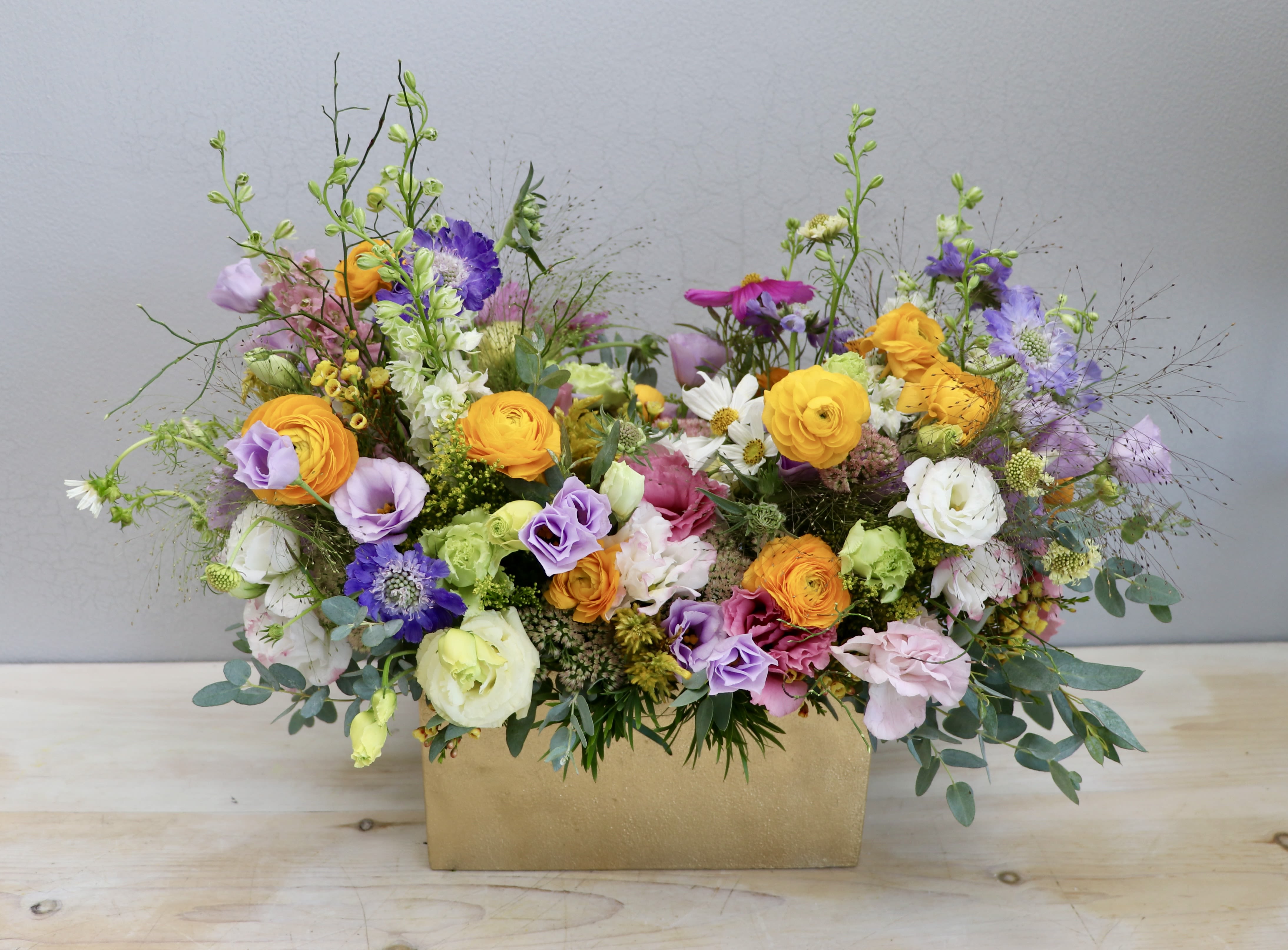 Colorful mixed flower arrangement with yellow, pink, and purple blooms in a rectangular gold container.