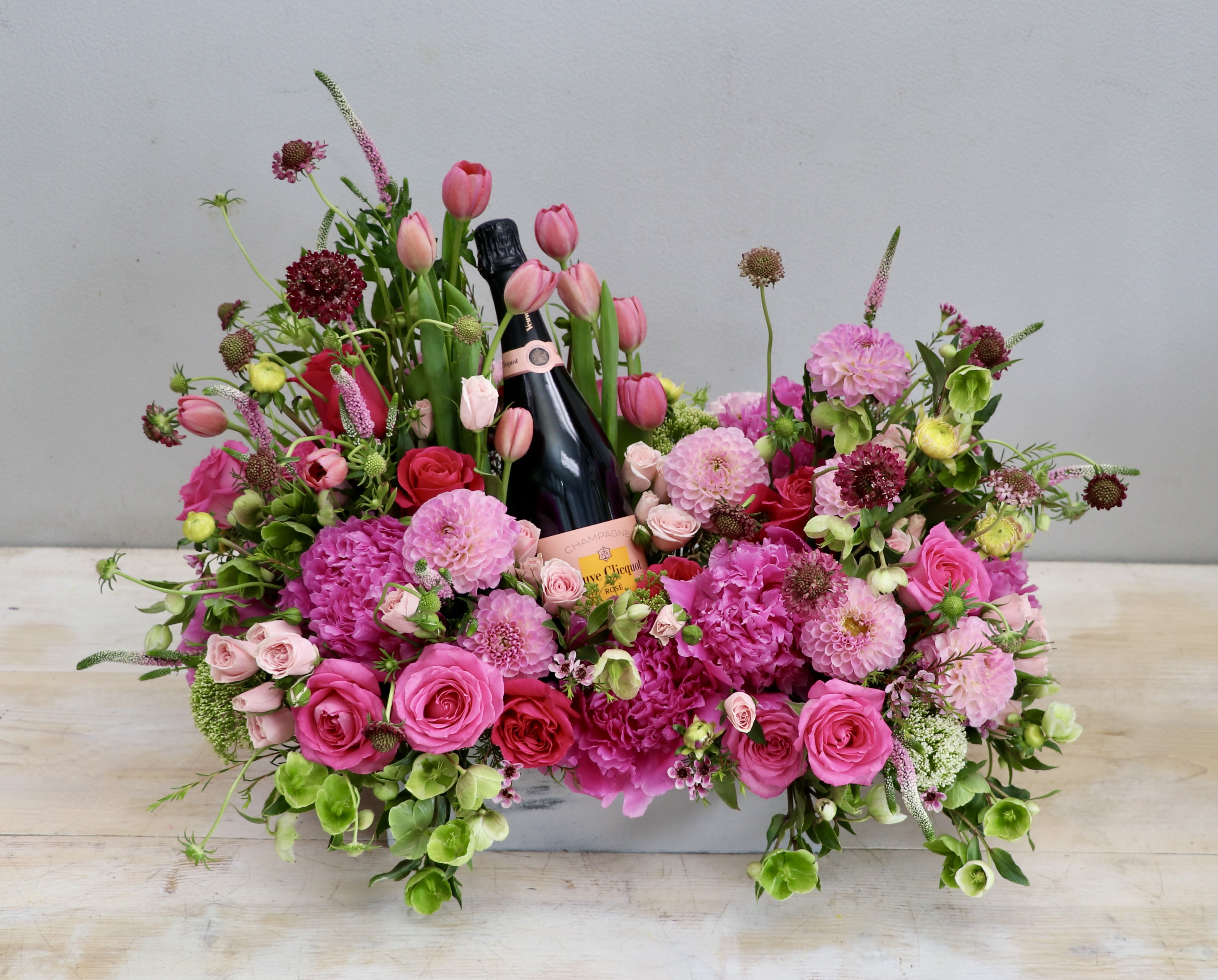 Low pink floral arrangement with roses and tulips surrounding a bottle of champagne in a white container.