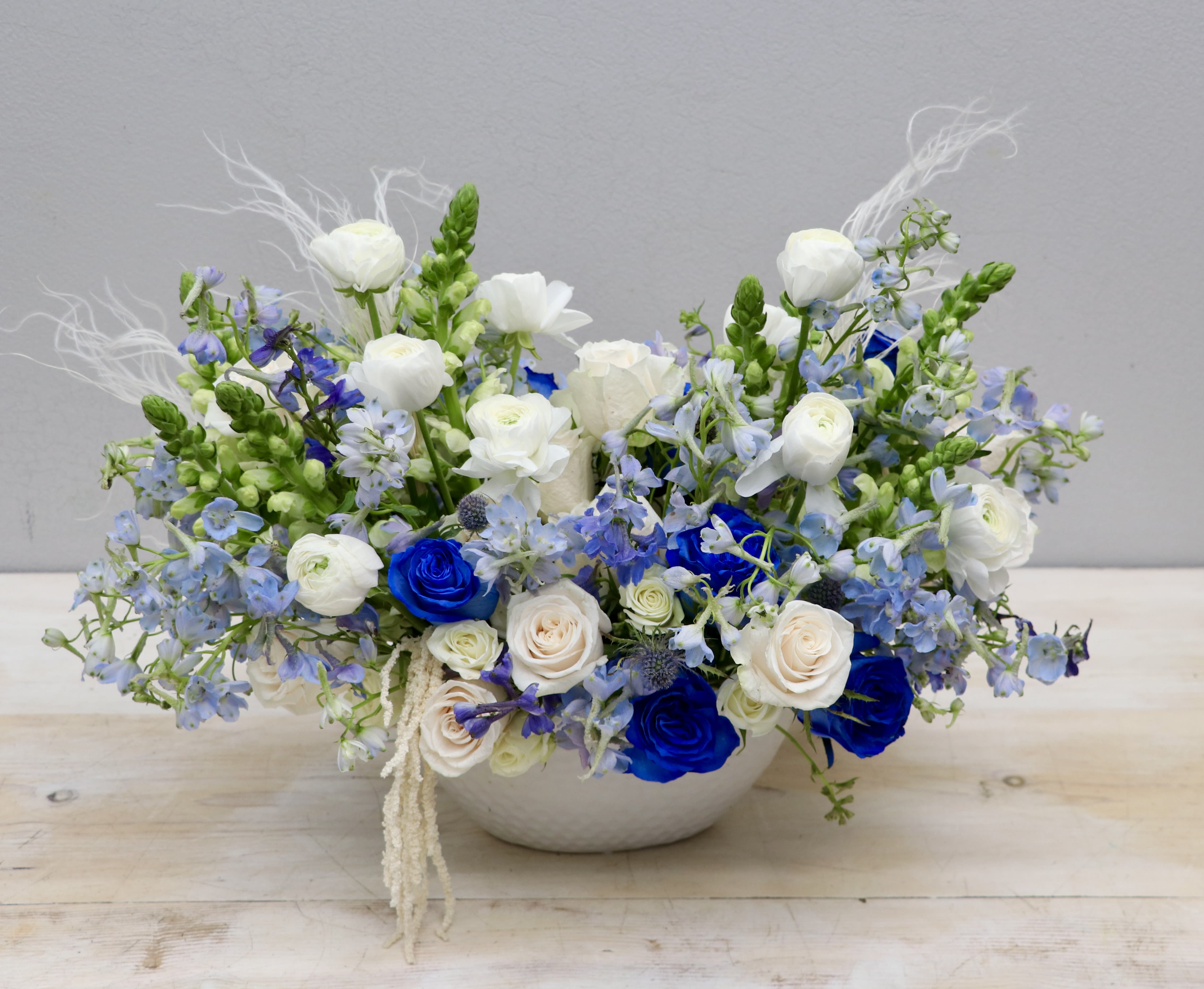 Low white bowl arrangement of blue and white roses with white ranunculus and light blue flowers
