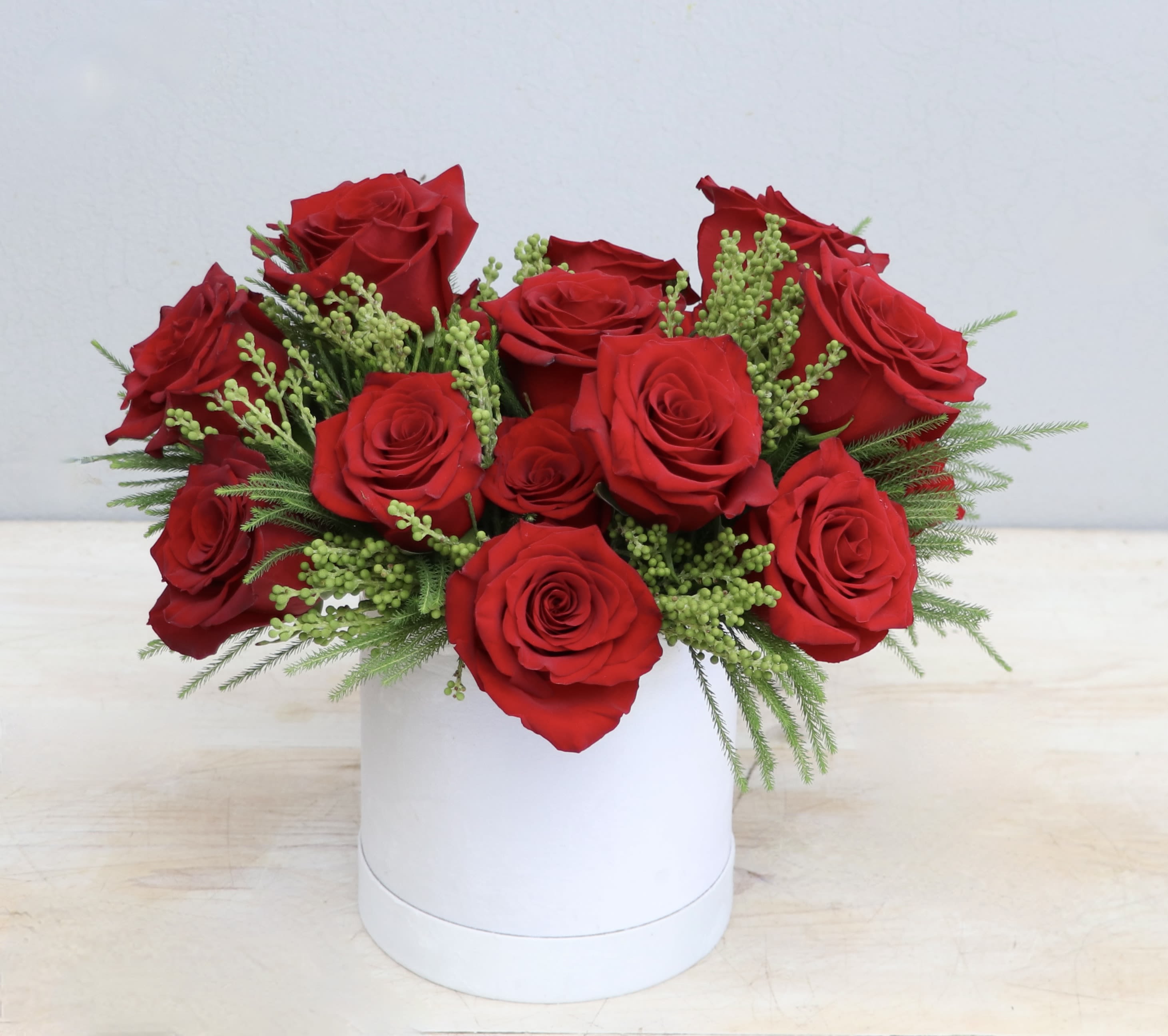 Low arrangement of red roses with green filler sprigs in a white round box.