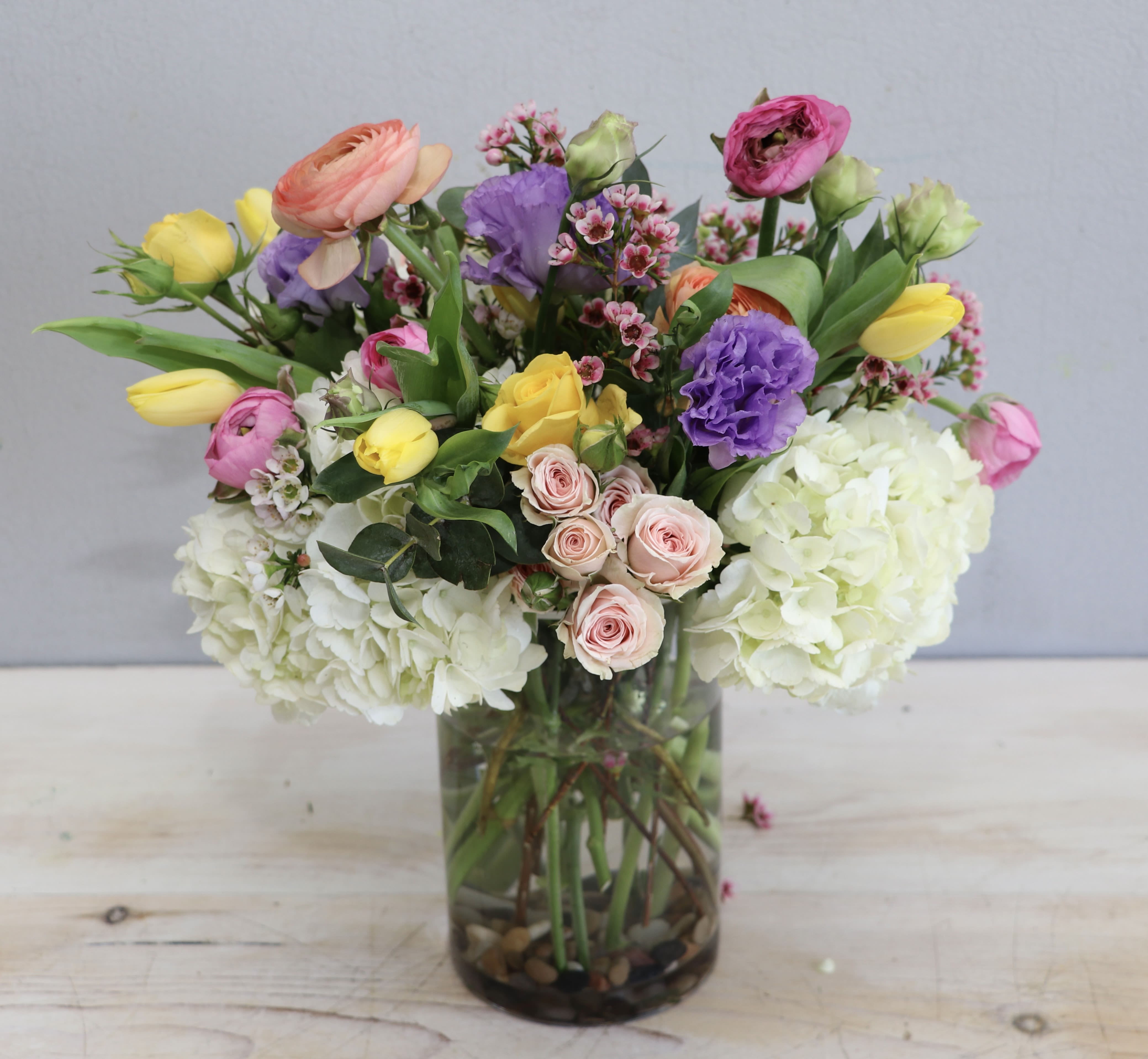 Spring Hues - Glendale Flowers - This sweet arrangement showcases a mix of ranunculus, tulips, spray roses and seasonal filler flowers with greenery. Standing at about 12-14 inches tall and wide at the standard size. 