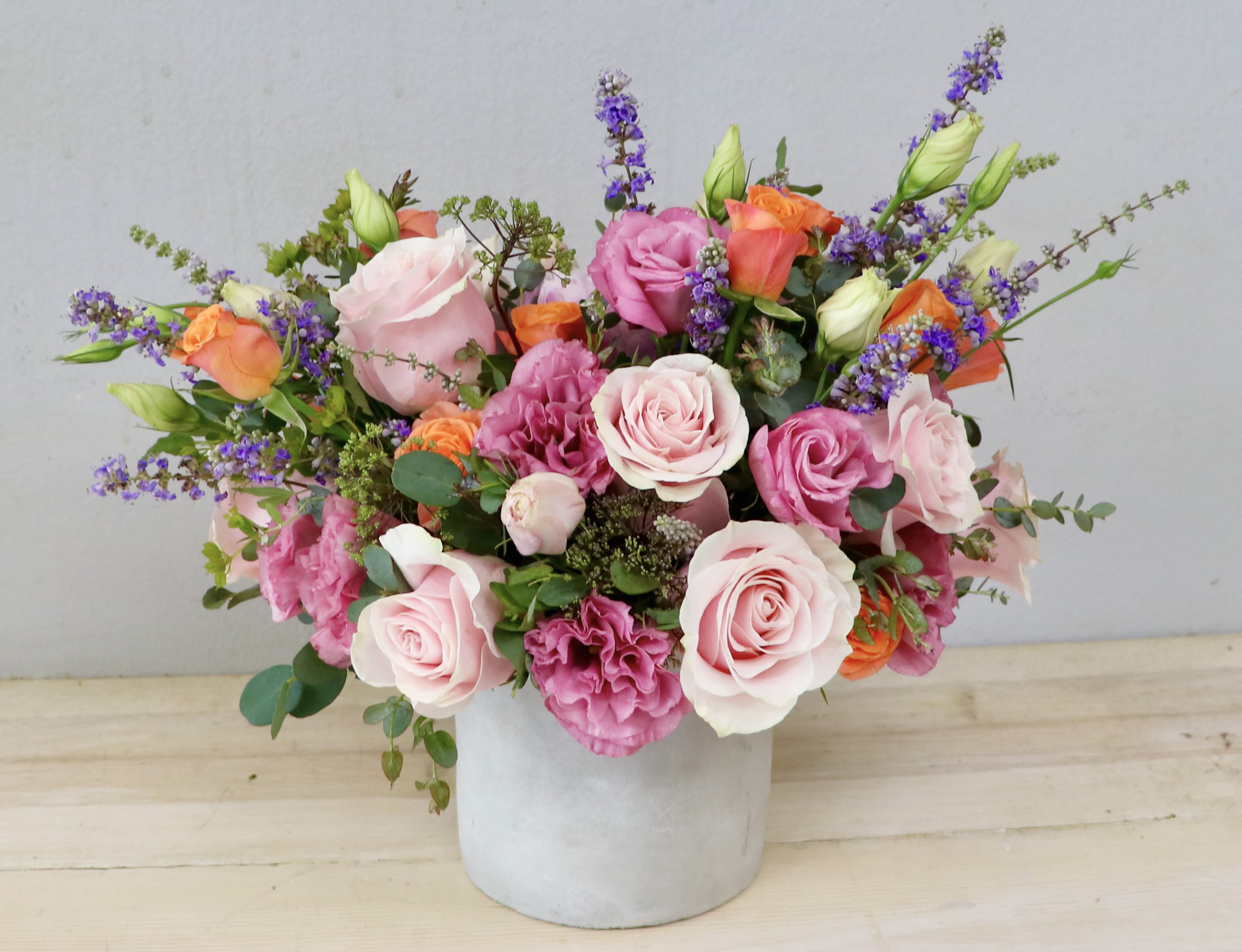 Whimsical Meadow - Glendale Flowers - Brimming with the effortless charm of a hidden cottage garden, this arrangement balances romantic elegance with a playful, vibrant energy. A lush foundation of premium garden roses and ruffled lisianthus in shades of soft blush, dusty mauve, and rich berry is beautifully interrupted by sudden, cheerful pops of bright orange spray roses.