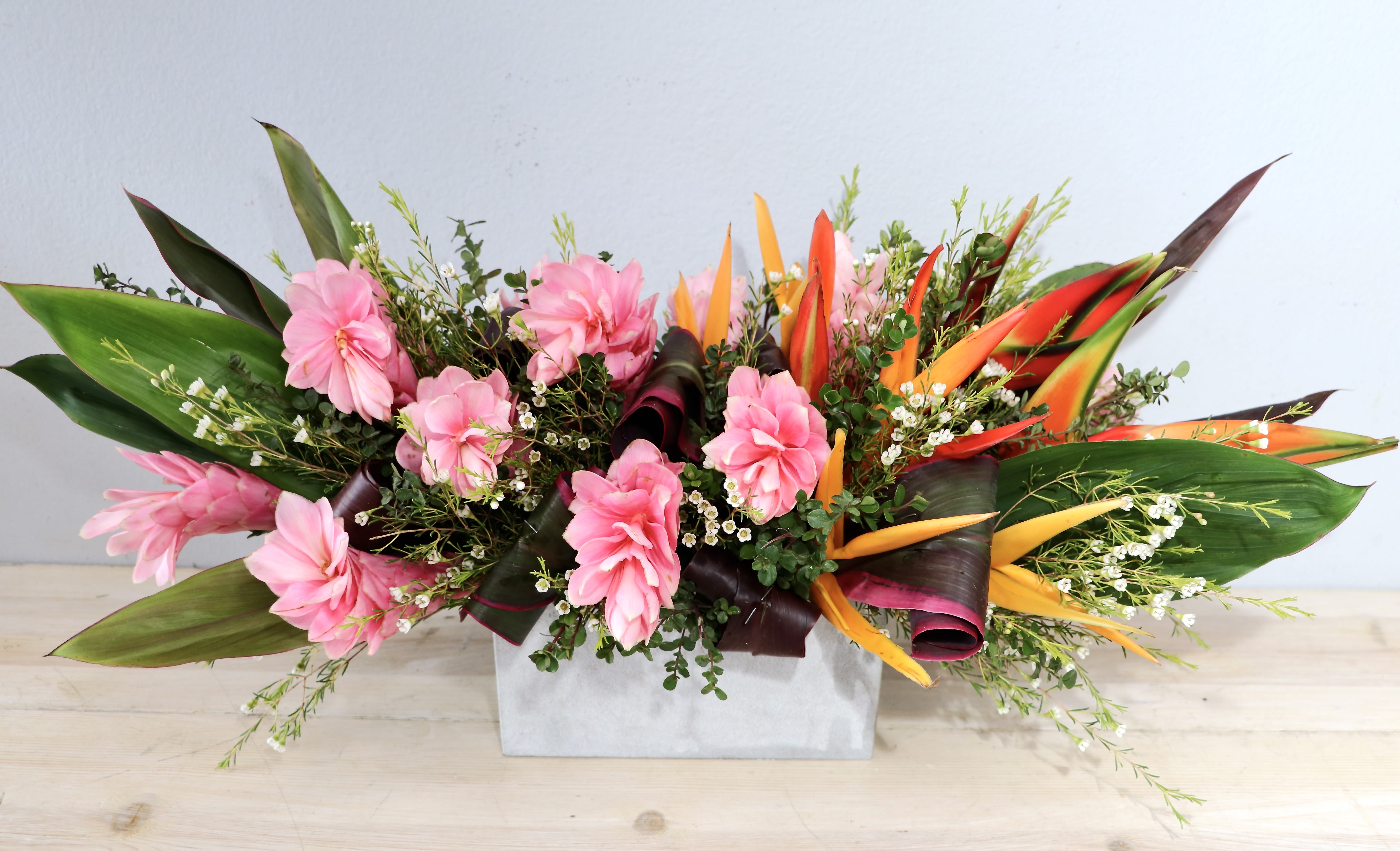 Ginger Tropics - Glendale Flowers - Bright, celebratory and a little exotic - this Ginger Tropics flower arrangement is a vibrant choice for a birthday, congratulations, or a cheerful just-because gift. The low, wide composition and the rectangular box presentation give it a modern, easy-to-place silhouette that immediately reads as festive for the person you're thinking of.  Clusters of vivid orange and red tropical blooms, set against broad dark green leaves and delicate white filler flowers for contrast and texture. The compact design keeps everything low and lush, making it ideal for tabletops, desks, or as a centerpiece that won't block conversation.  Designed to make someone feel seen and celebrated, this arrangement delivers bold color and tropical energy without being overpowering. Care is simple-keep the floral foam hydrated and display in a cool spot away from direct sunlight to extend freshness.
