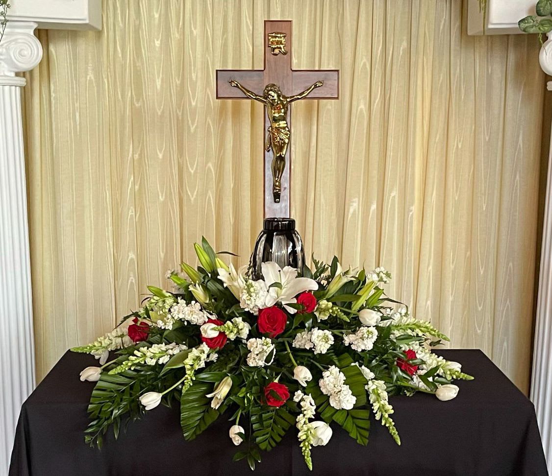 Sympathy floral arrangement with red roses, white lilies, and a crucifix backdrop
