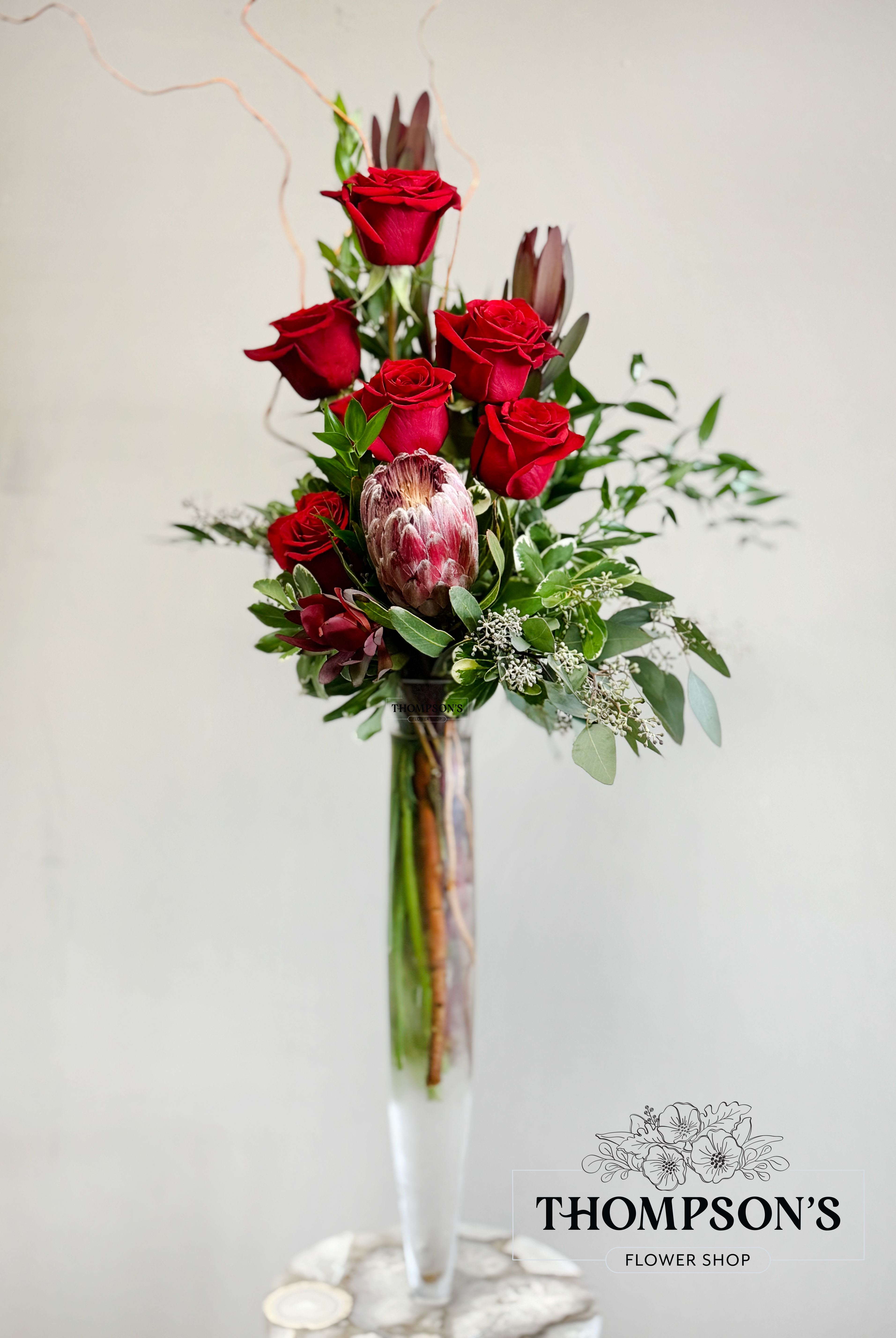 Tall vase arrangement of red roses with greenery and a protea
