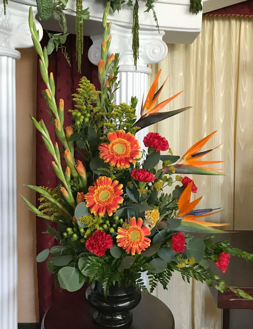 Tall arrangement of orange gerberas, red carnations, and birds of paradise in a black urn vase