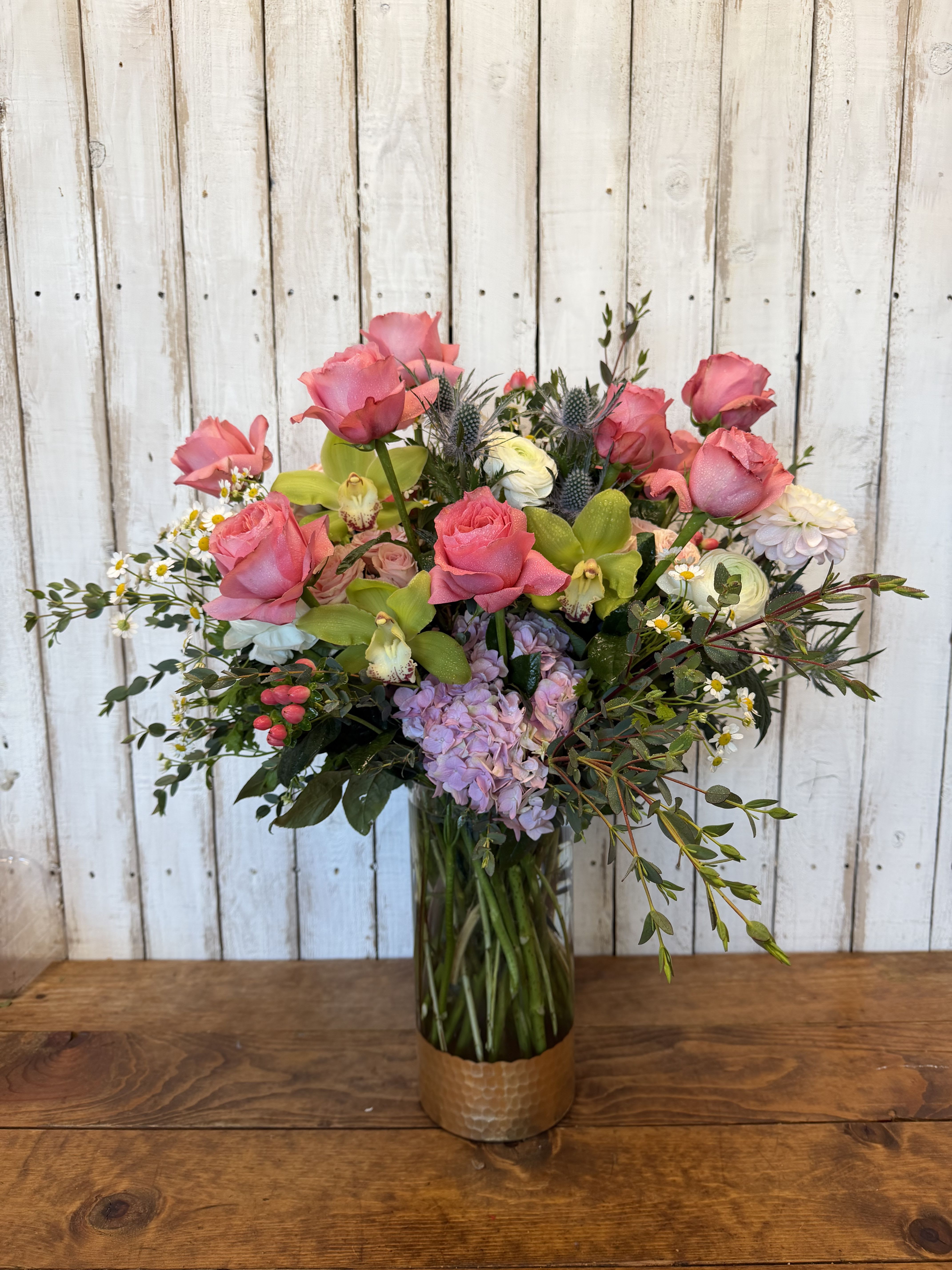 Bridgerton‘s Garden - A dreamy garden-style arrangement designed in a clear glass vase with a gold-dipped base, featuring lush garden roses, delicate orchids, and soft snapdragons accented with airy filler flowers. This design has a romantic, whimsical look that feels straight out of a Bridgerton garden party—timeless, elegant, and full of graceful movement.
