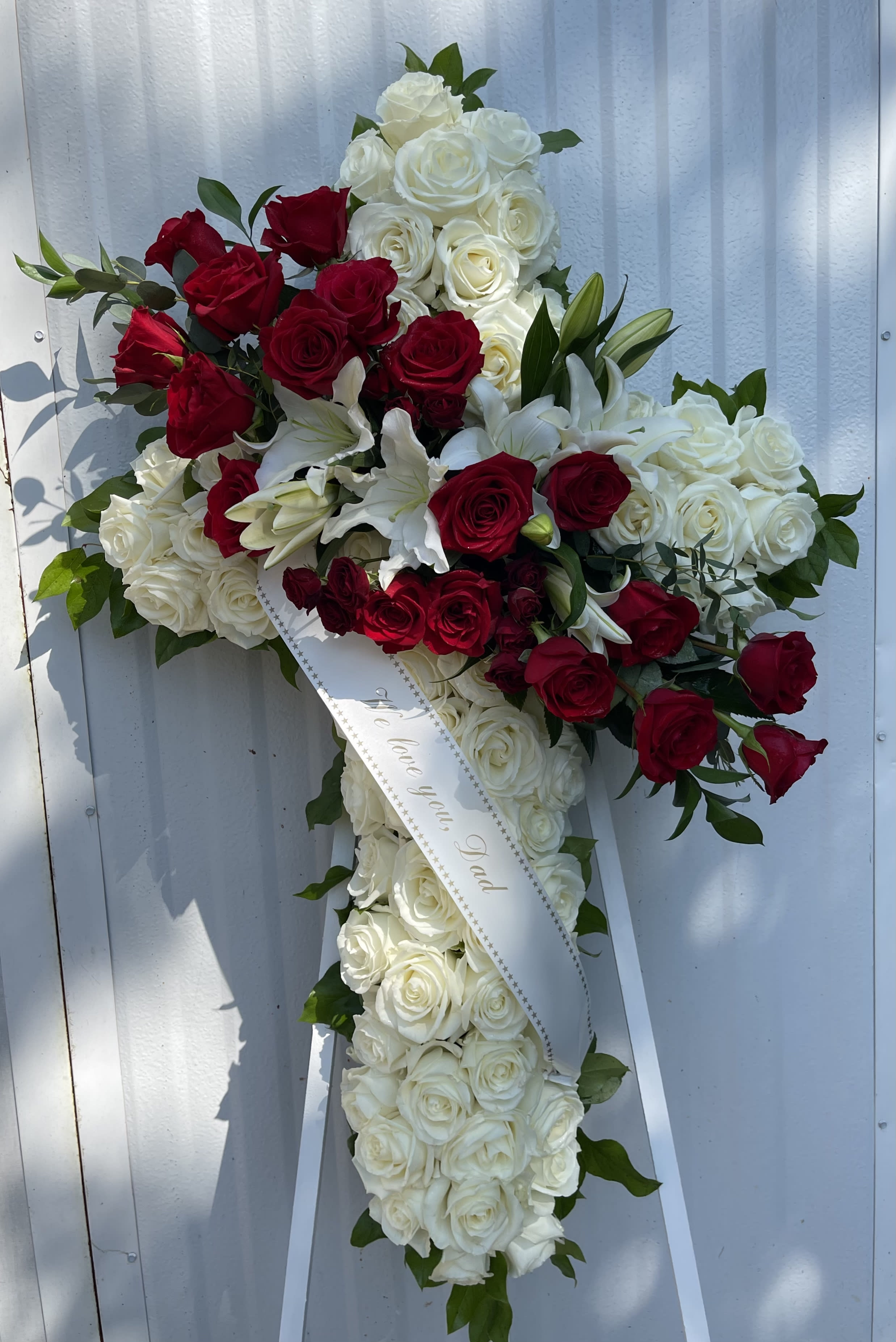 Cross-shaped floral spray of red and white roses with lilies and a memorial ribbon