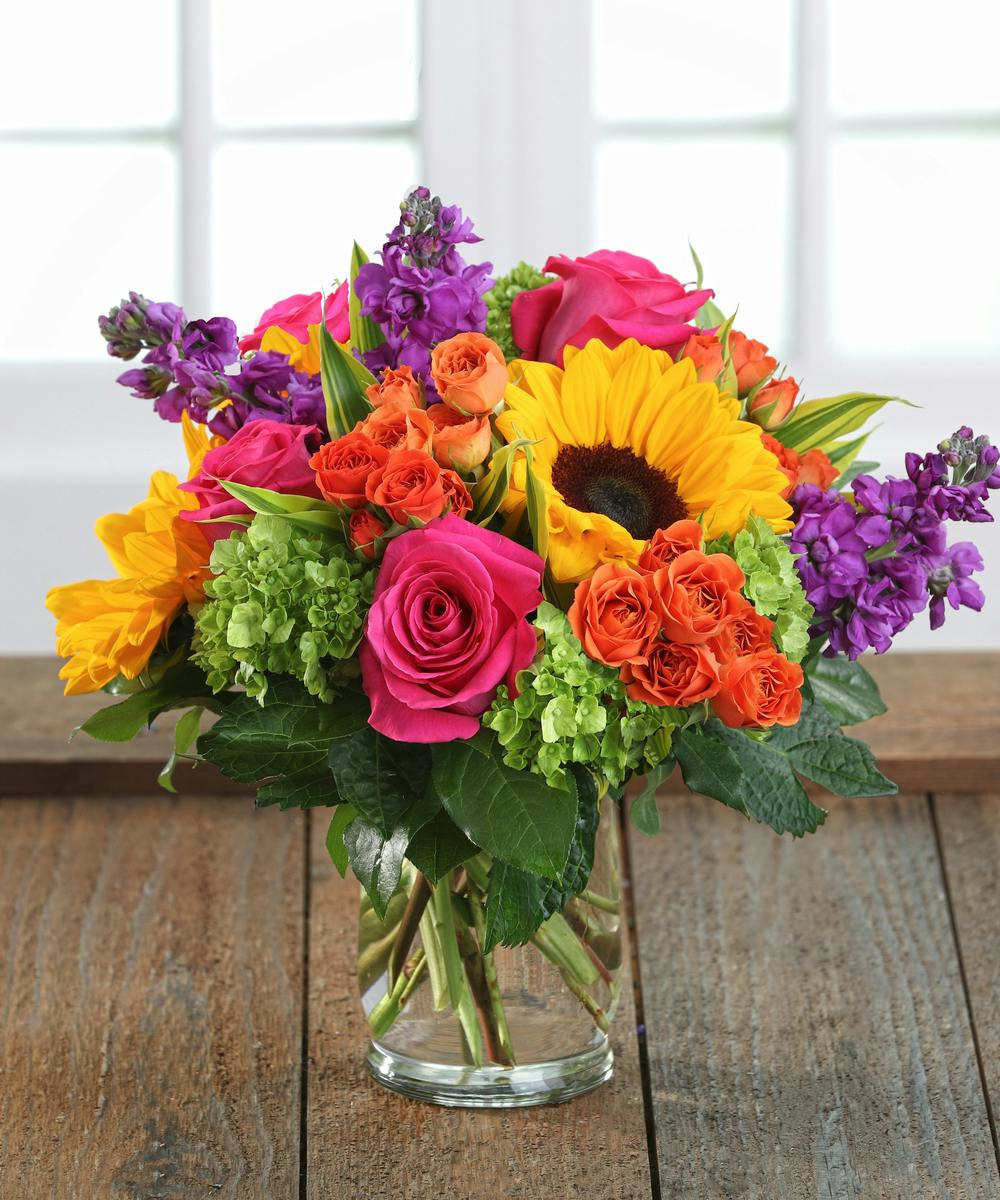 Bright mixed bouquet of roses, sunflowers, and purple blooms in a glass vase