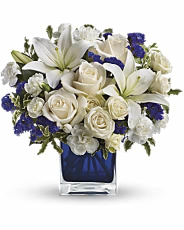 White lilies and roses with blue accents in a square glass vase