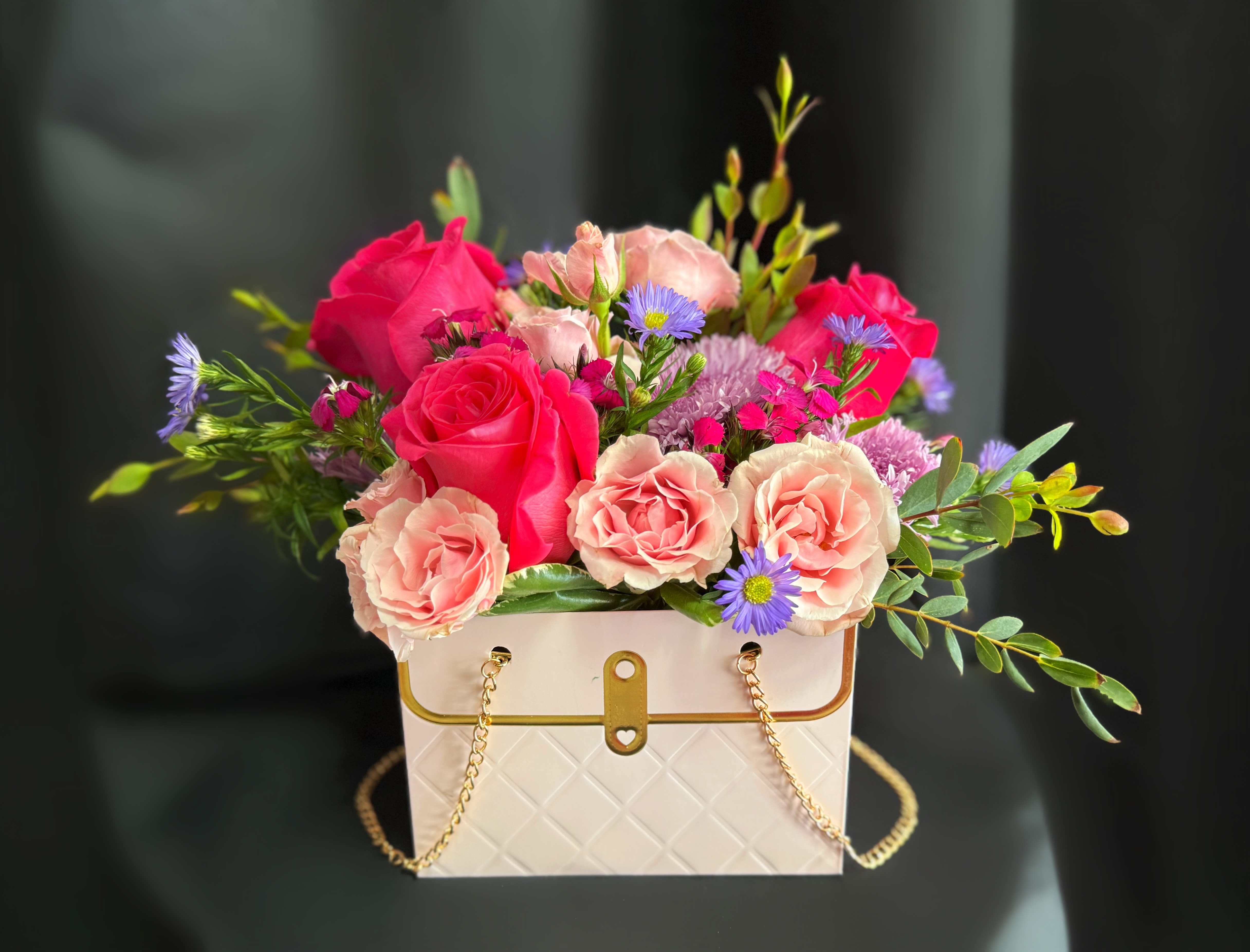 Pink roses and purple daisies arranged in a white handbag-shaped box
