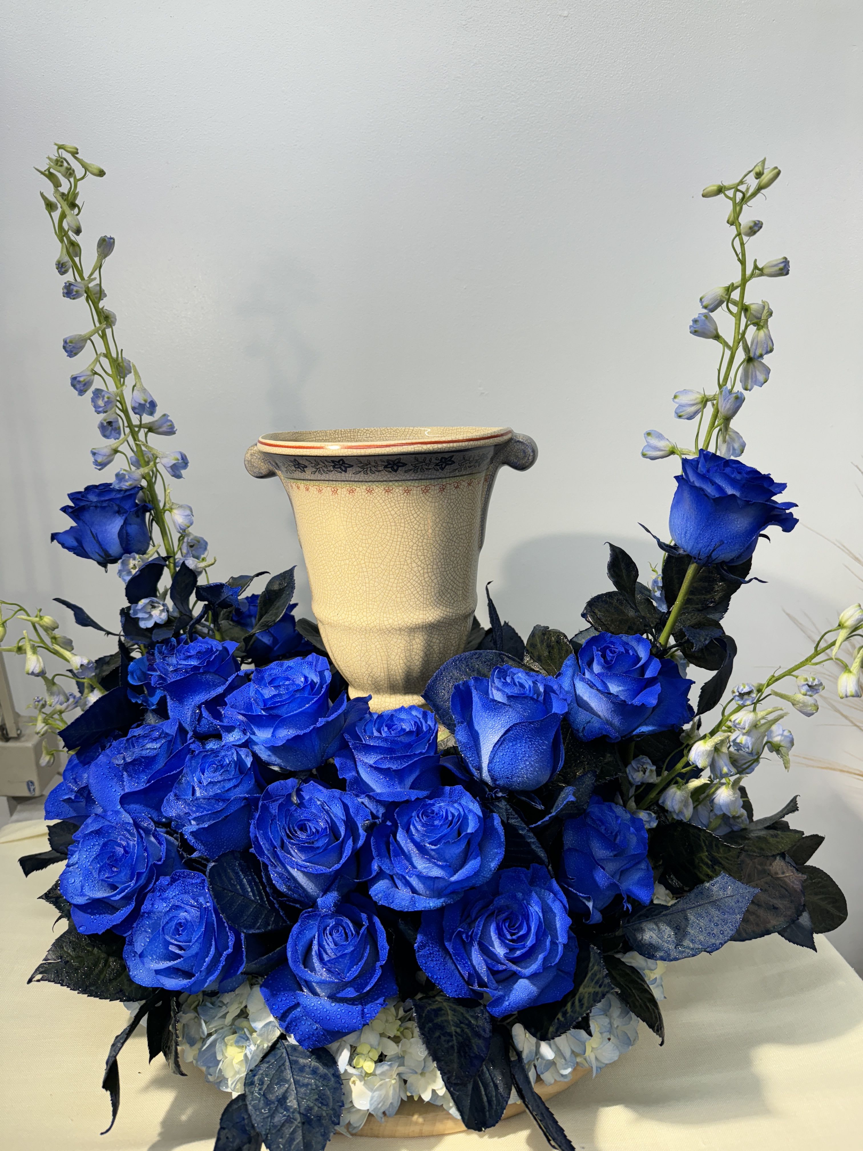 Blue roses arranged around a beige urn vase with tall blue flower spikes